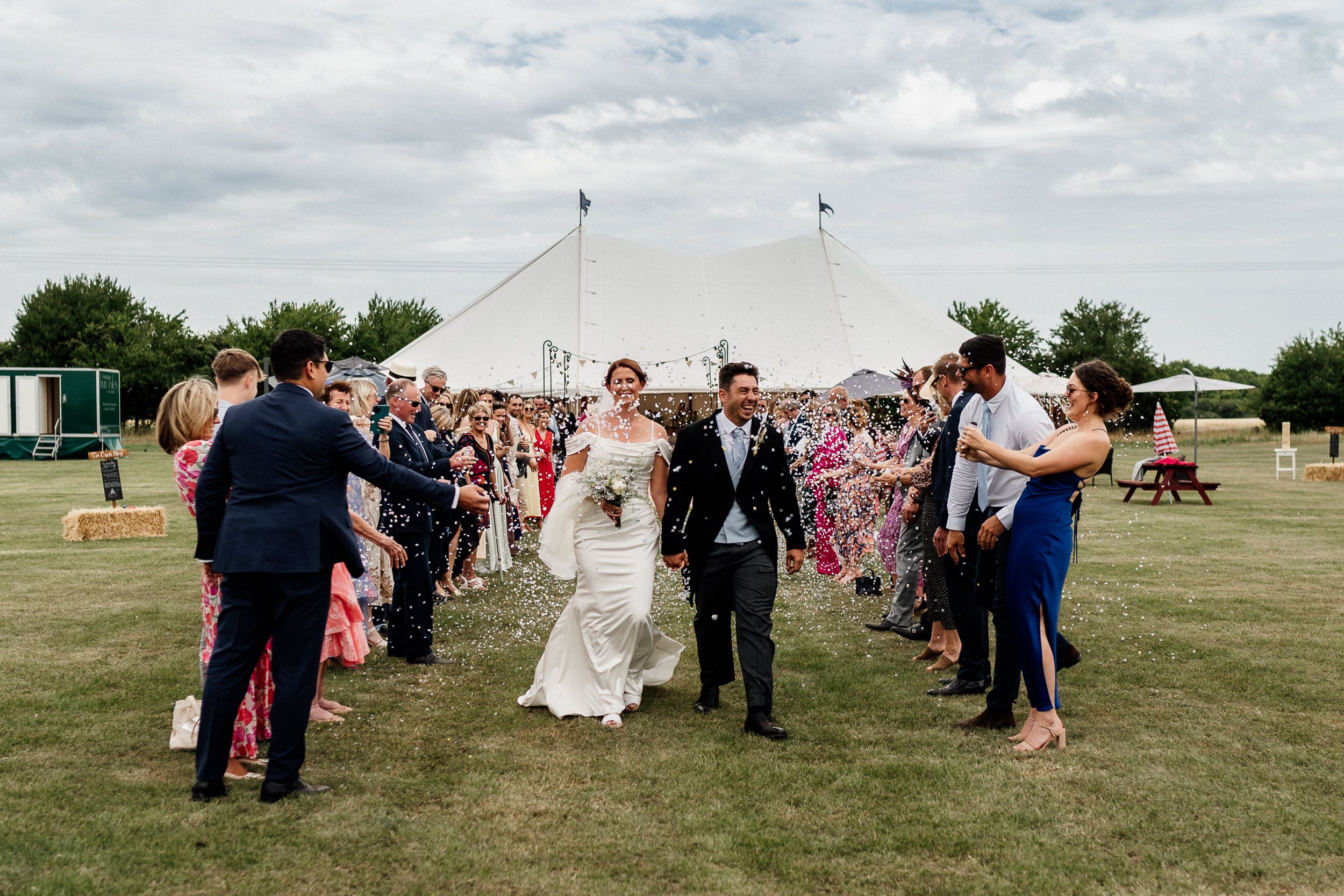 Wedding guests stand in a grassy field during an outdoor reception, forming a line under cloudy skies.