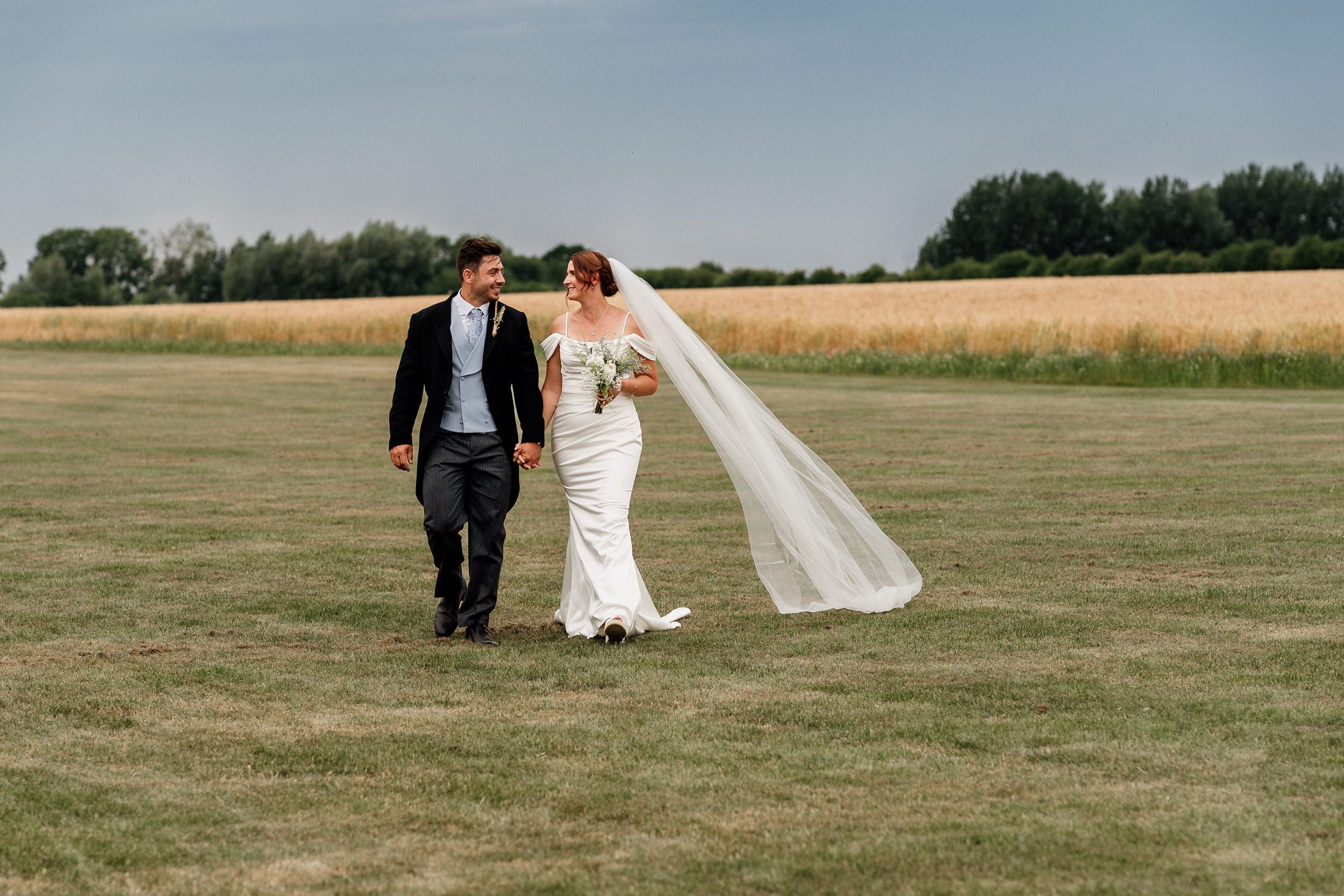 Bride and groom walk hand in hand through an open summer field as her white veil flows in the breeze.