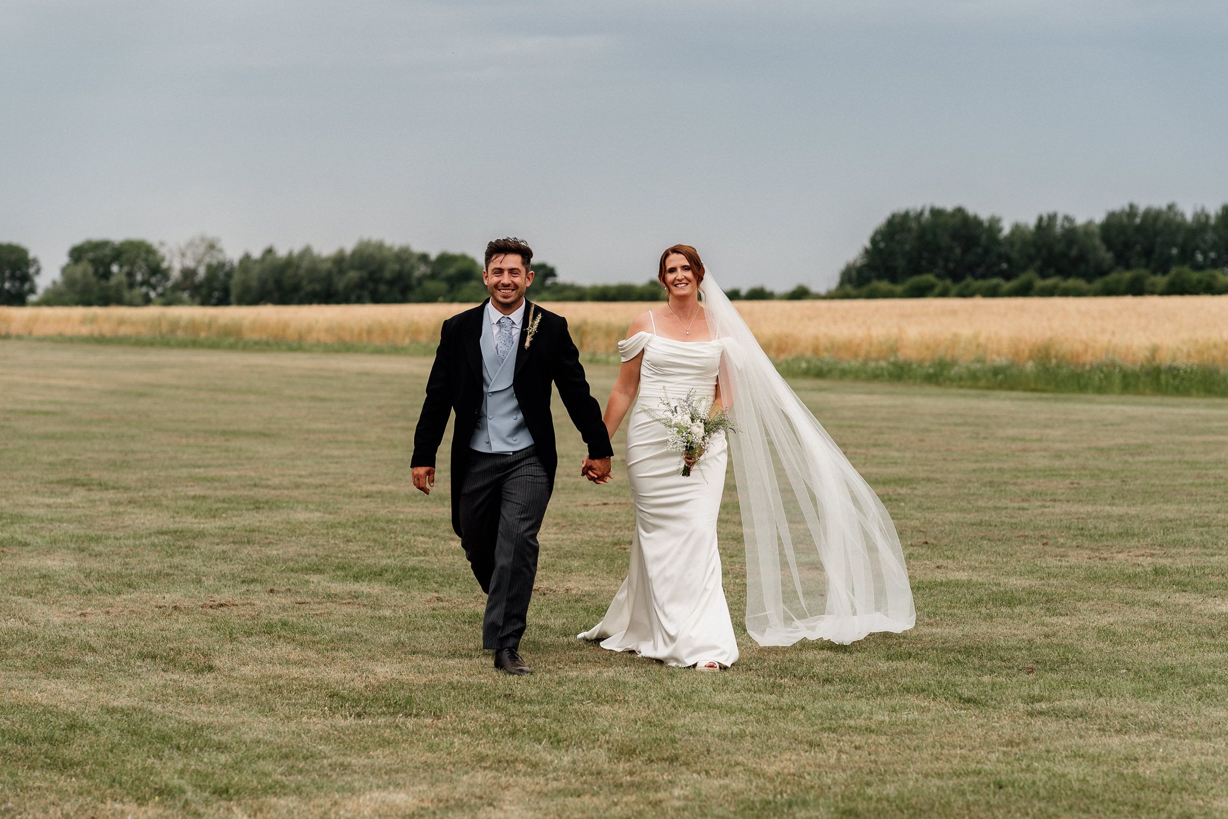 A couple walks hand in hand across a grassy field with a long veil flowing in the wind.