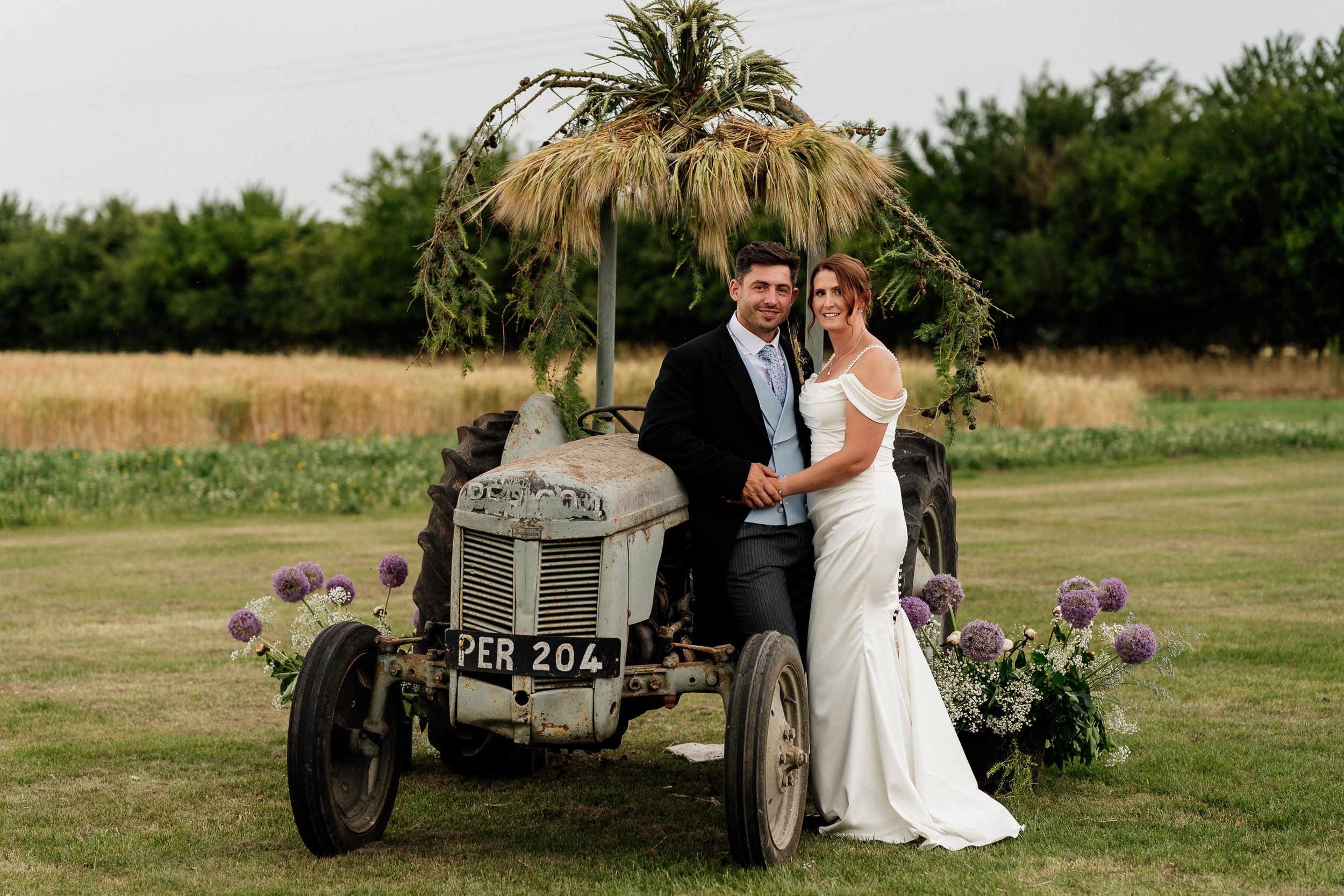 Couple in formal wedding attire poses in rustic outdoor countryside setting near vintage tractor and green fields.