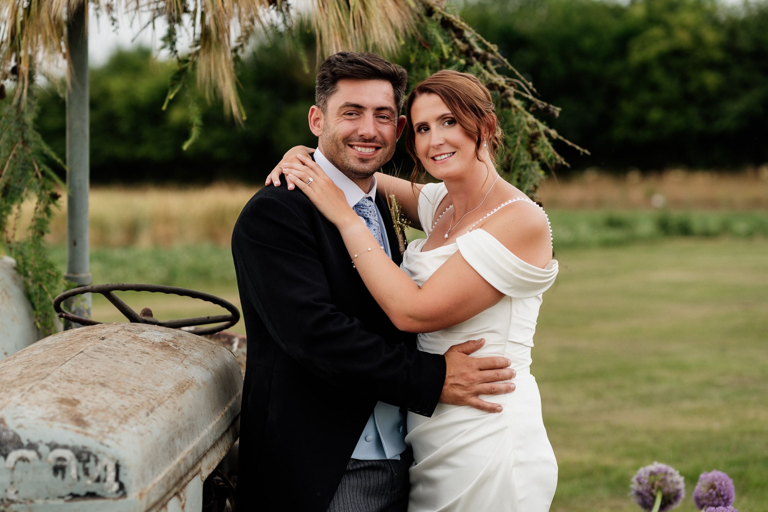 Happy newlywed couple embraces beside vintage tractor on their wedding day in rustic outdoor setting.