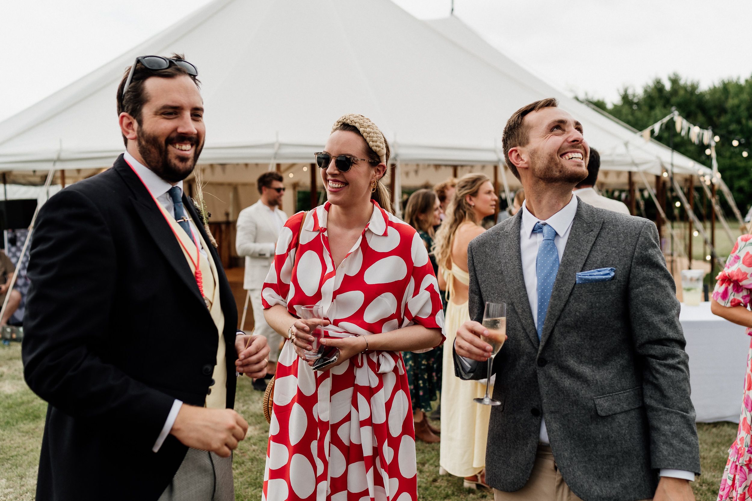 Three guests share a laugh during an outdoor wedding reception with a white tent in the background.