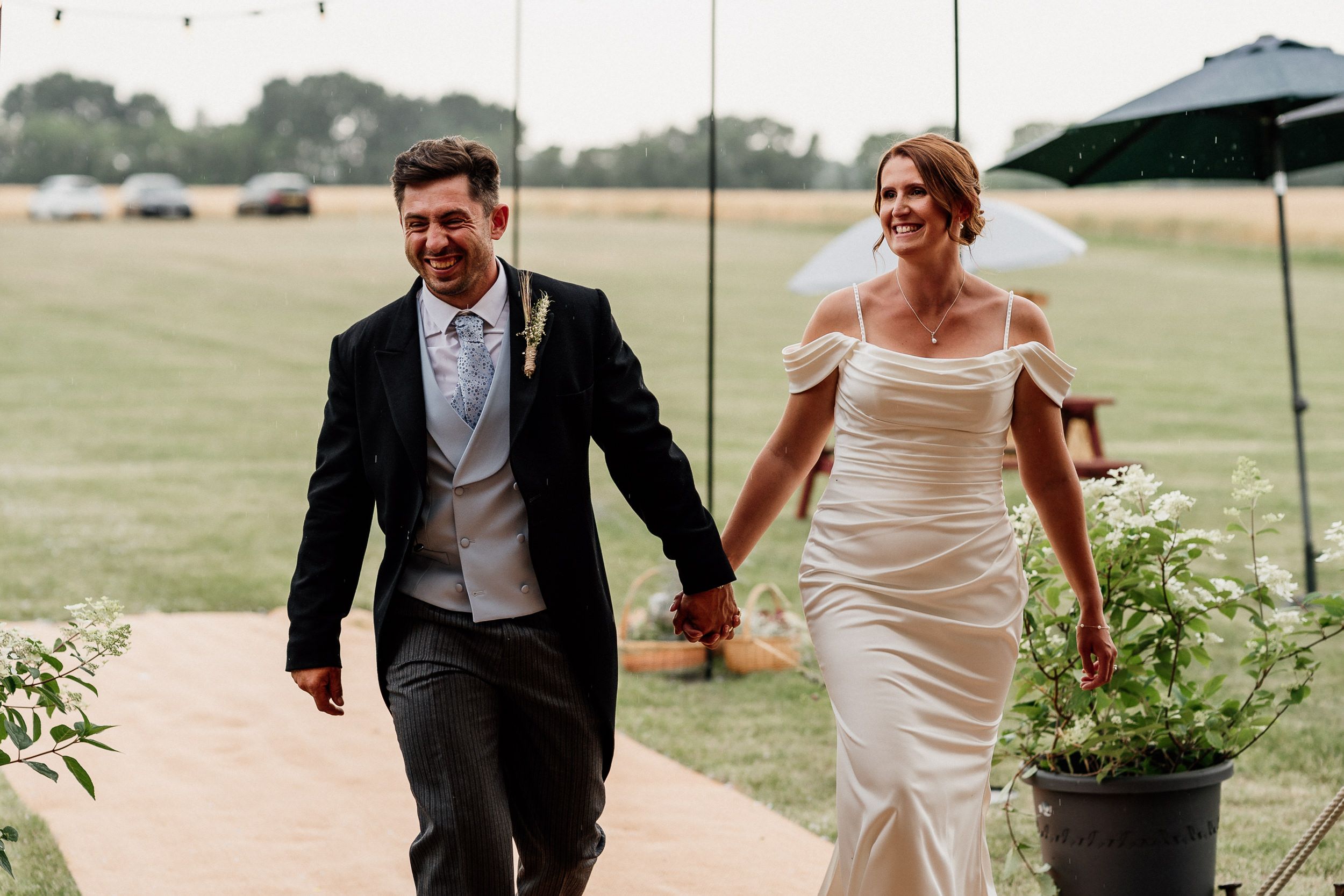 A bride and groom run playfully through a grassy field during their outdoor wedding celebration.