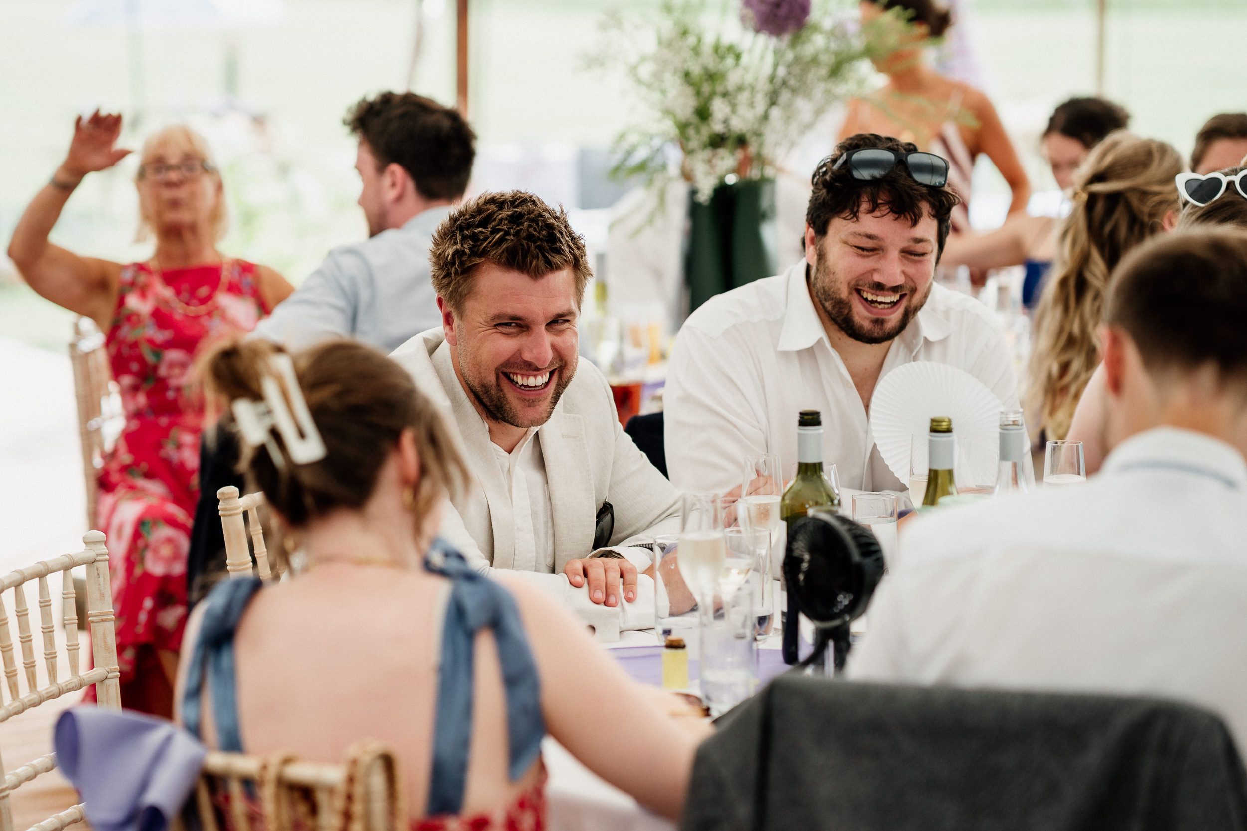Wedding guests laugh and celebrate together at a reception table inside an elegant tent with lively conversation.