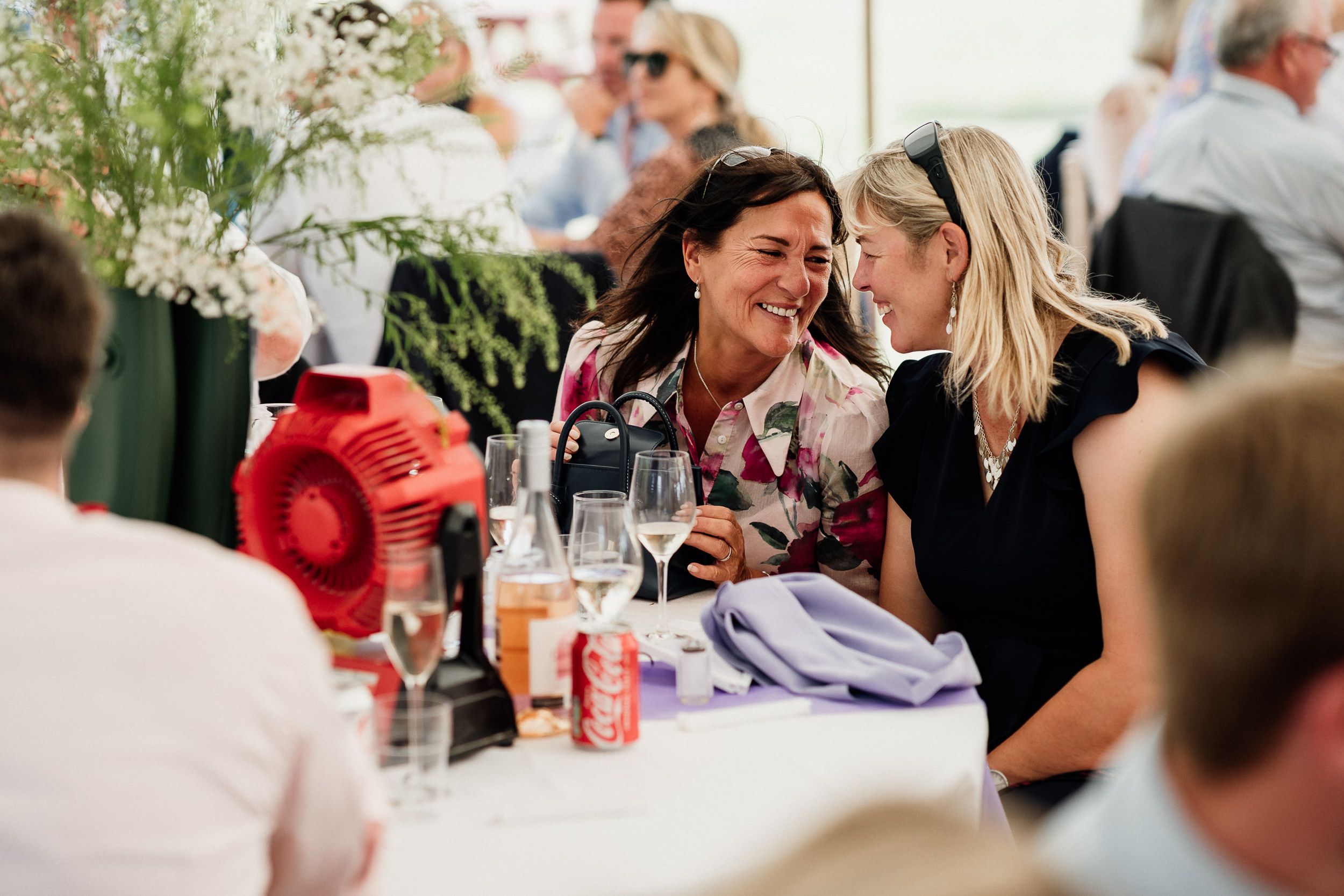 Two people laugh and chat together at an outdoor dining table with wine glasses and floral centerpieces.