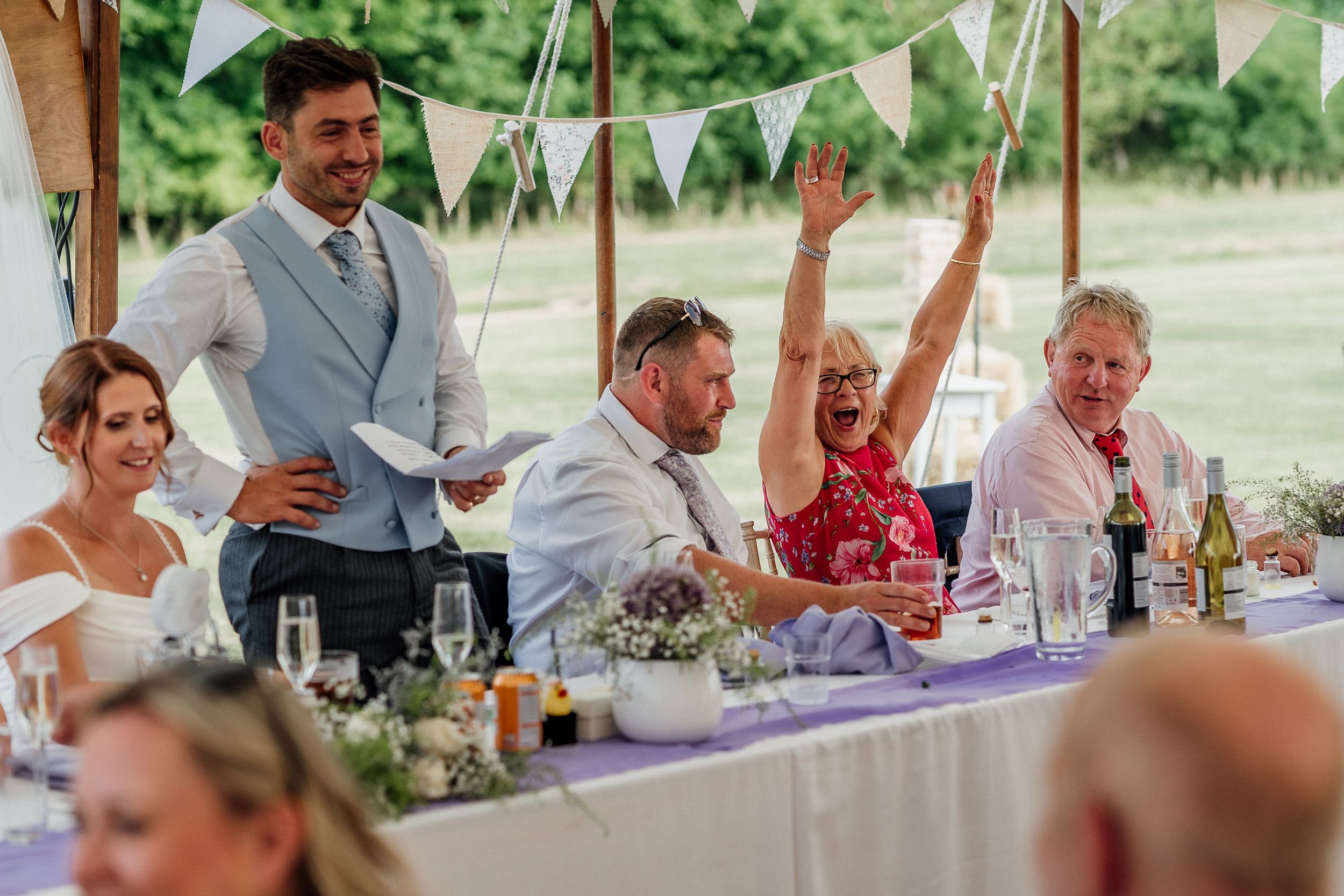 Wedding party celebrates with raised arms during speeches at a festive reception decorated with bunting and florals.