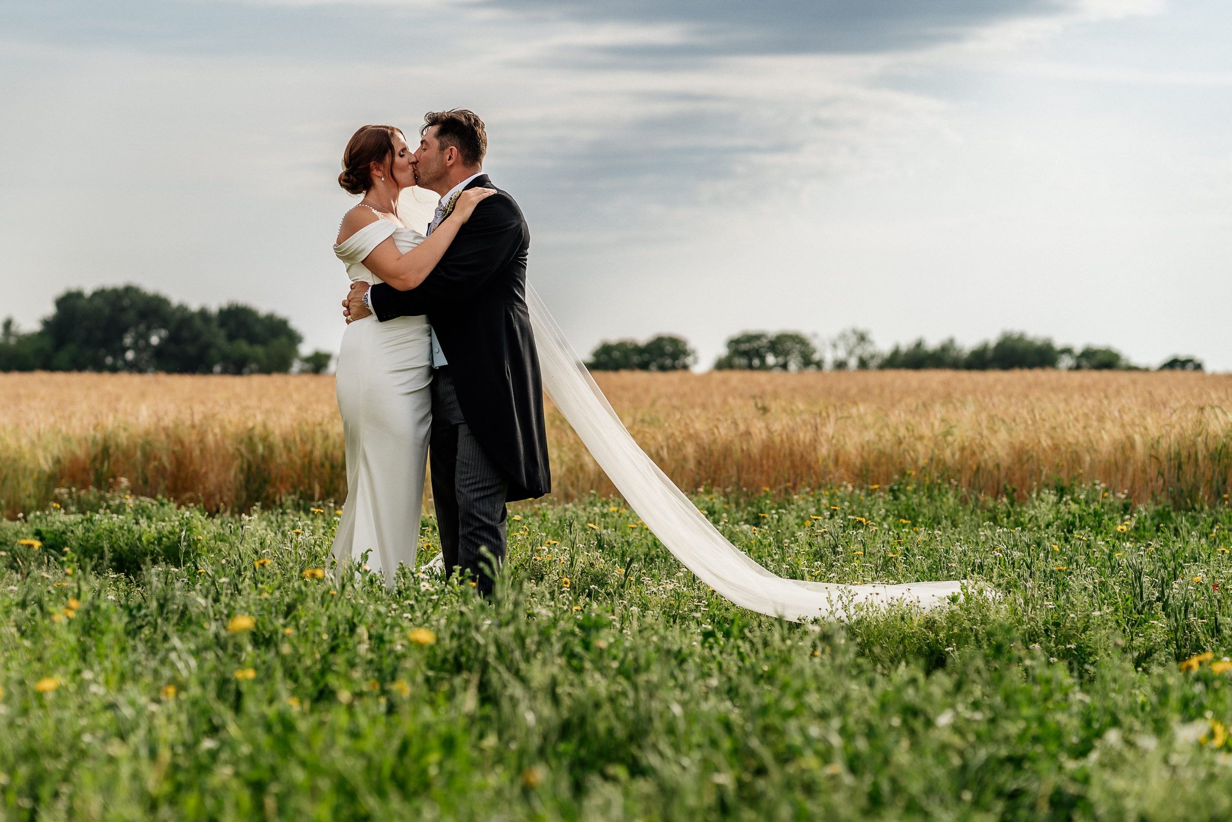 A couple shares a romantic moment in a beautiful field at sunset, with a flowing white wedding dress train trailing behind.