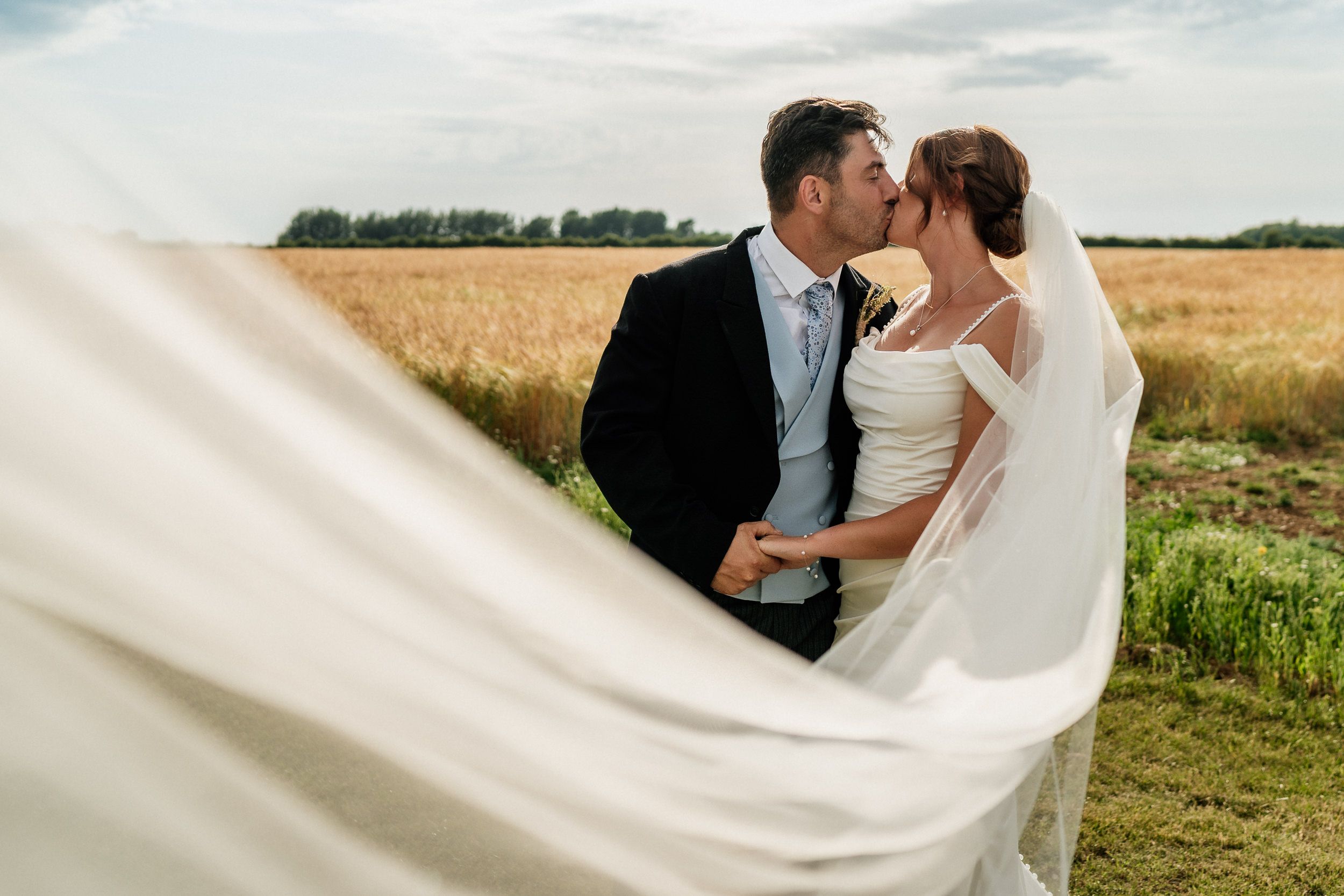 A couple shares a romantic kiss in a wheat field while the bride's white veil flows dramatically in the wind.