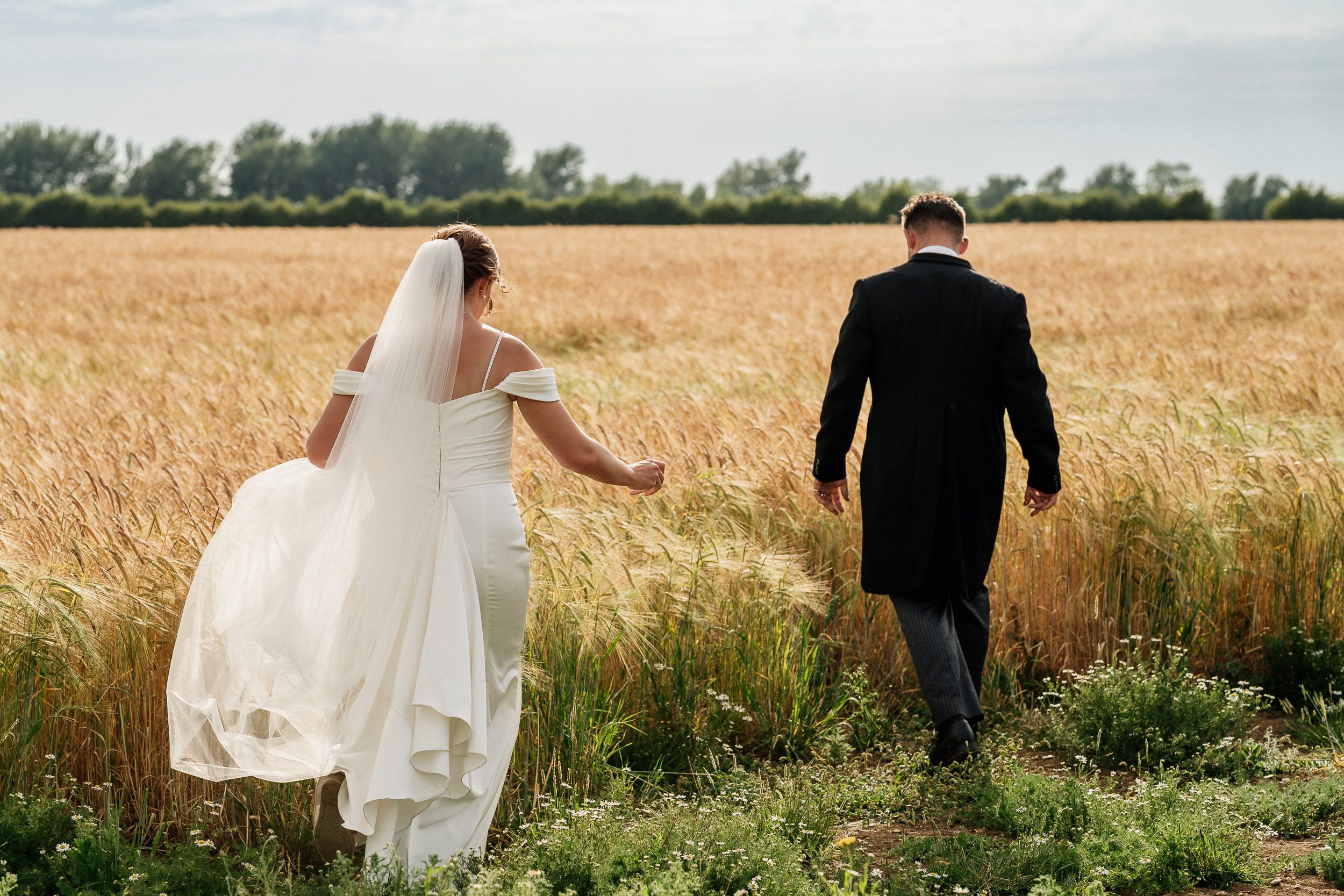 A bride and groom walk hand in hand through a golden wheat field on their wedding day.