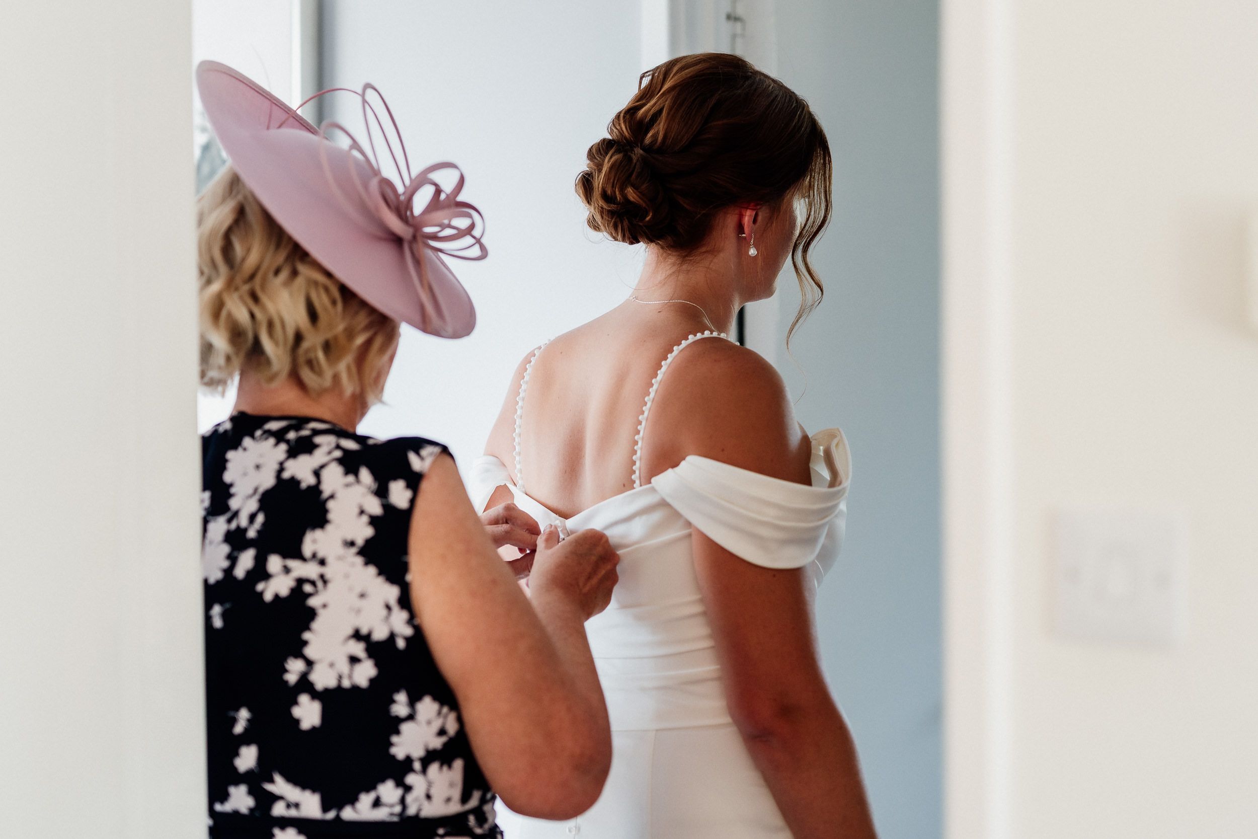 Someone helps adjust the back of an elegant white off-shoulder wedding dress as the bride prepares for her ceremony.