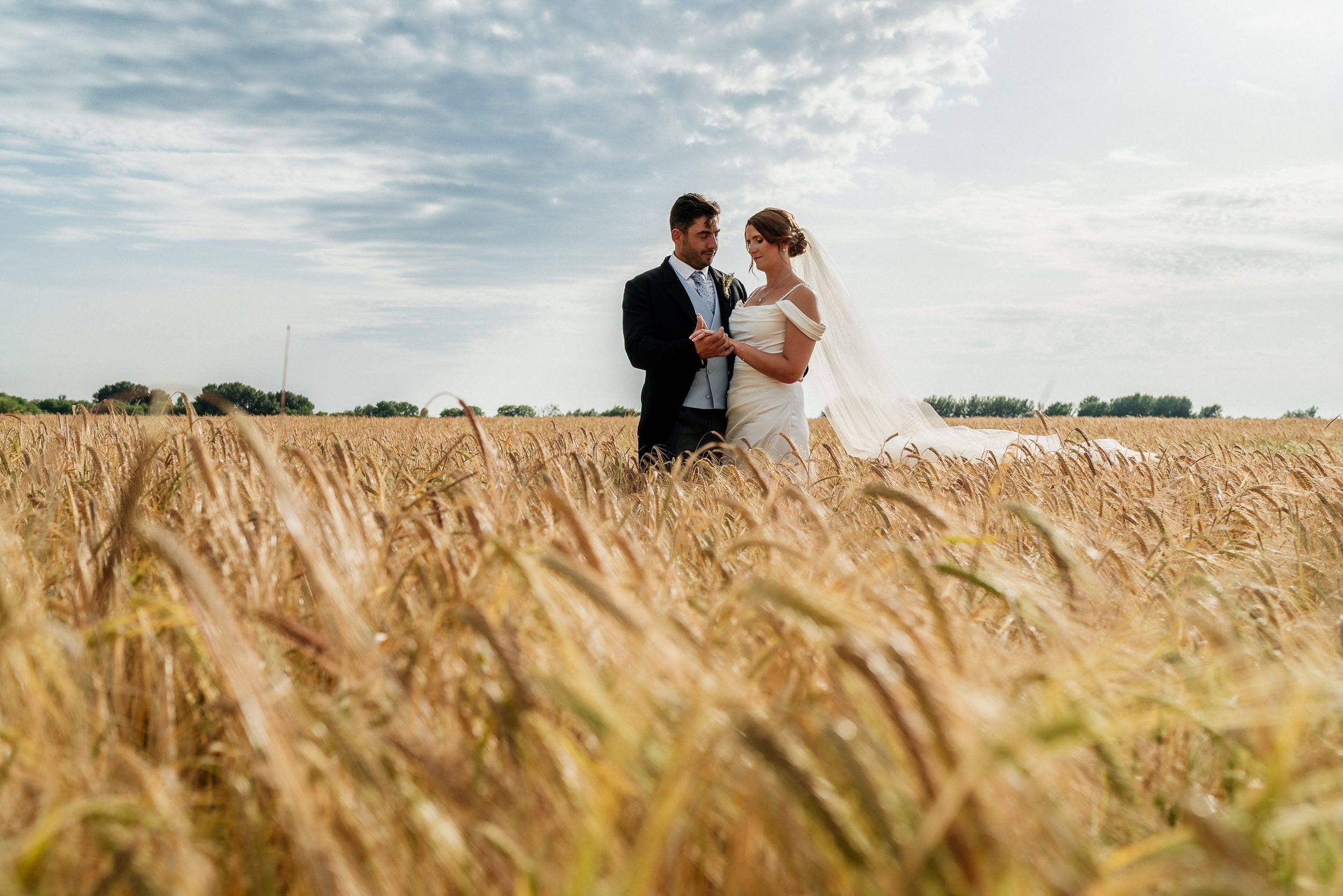 A bride and groom stand together in a golden wheat field under a cloudy summer sky.