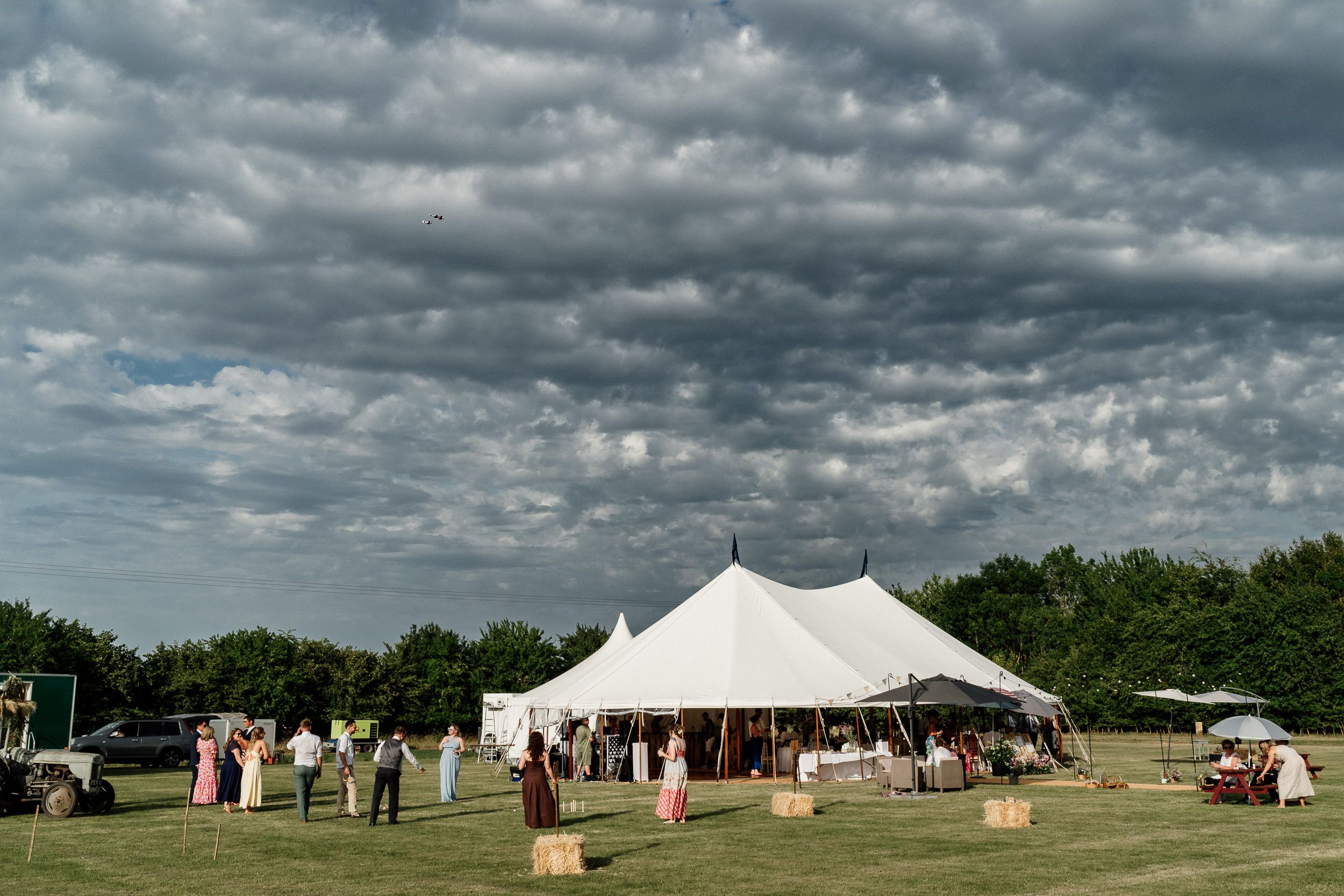 A panoramic view of white tents set up across a grassy field under dramatic cloudy skies on a summer day.