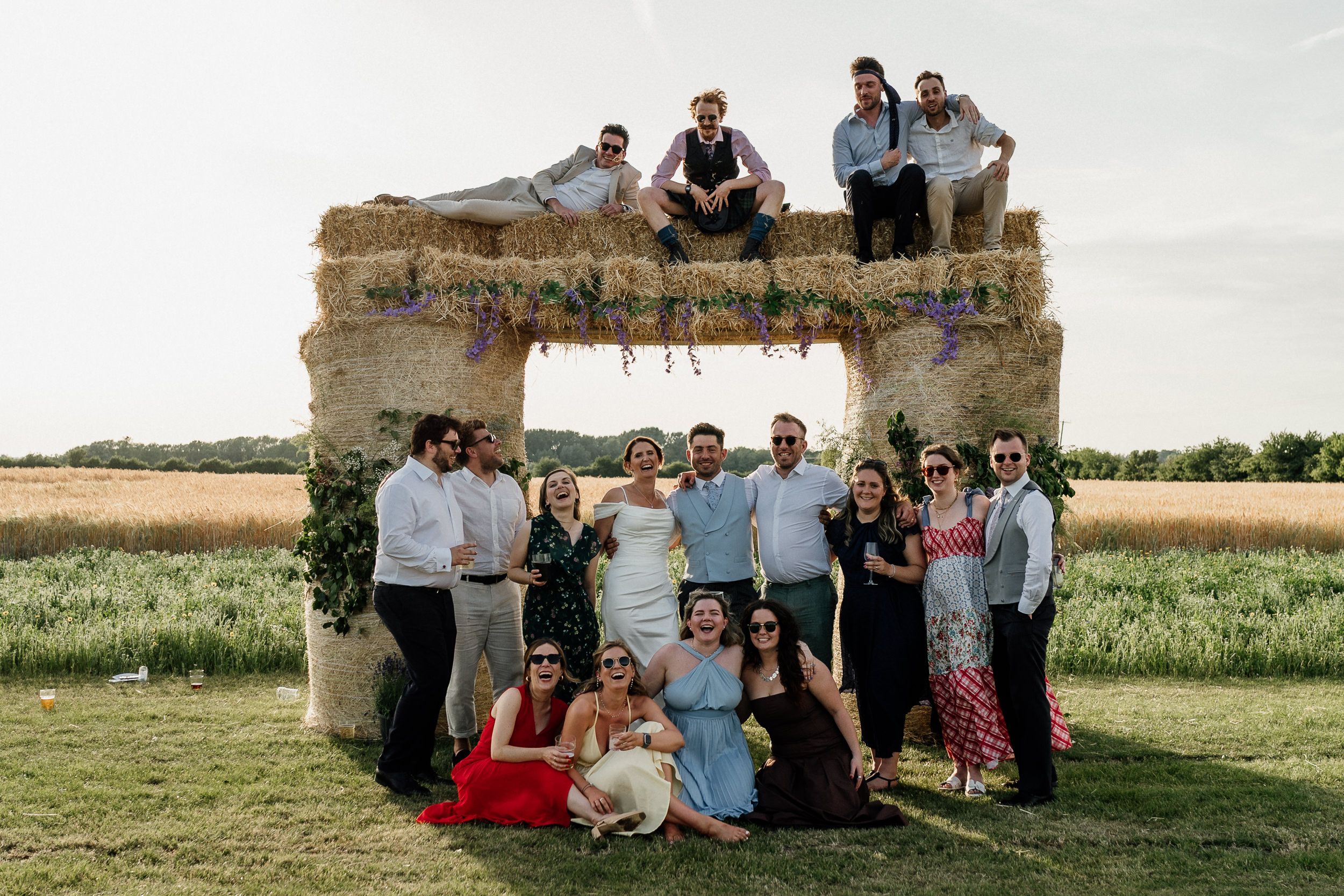 Wedding party poses together at rustic outdoor stone archway in countryside field during summer celebration.