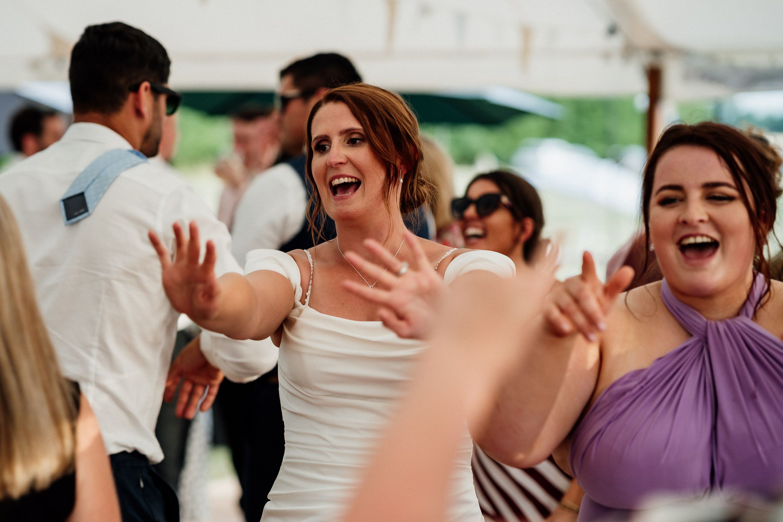 Wedding guests laugh and dance together during an outdoor celebration under a white tent.