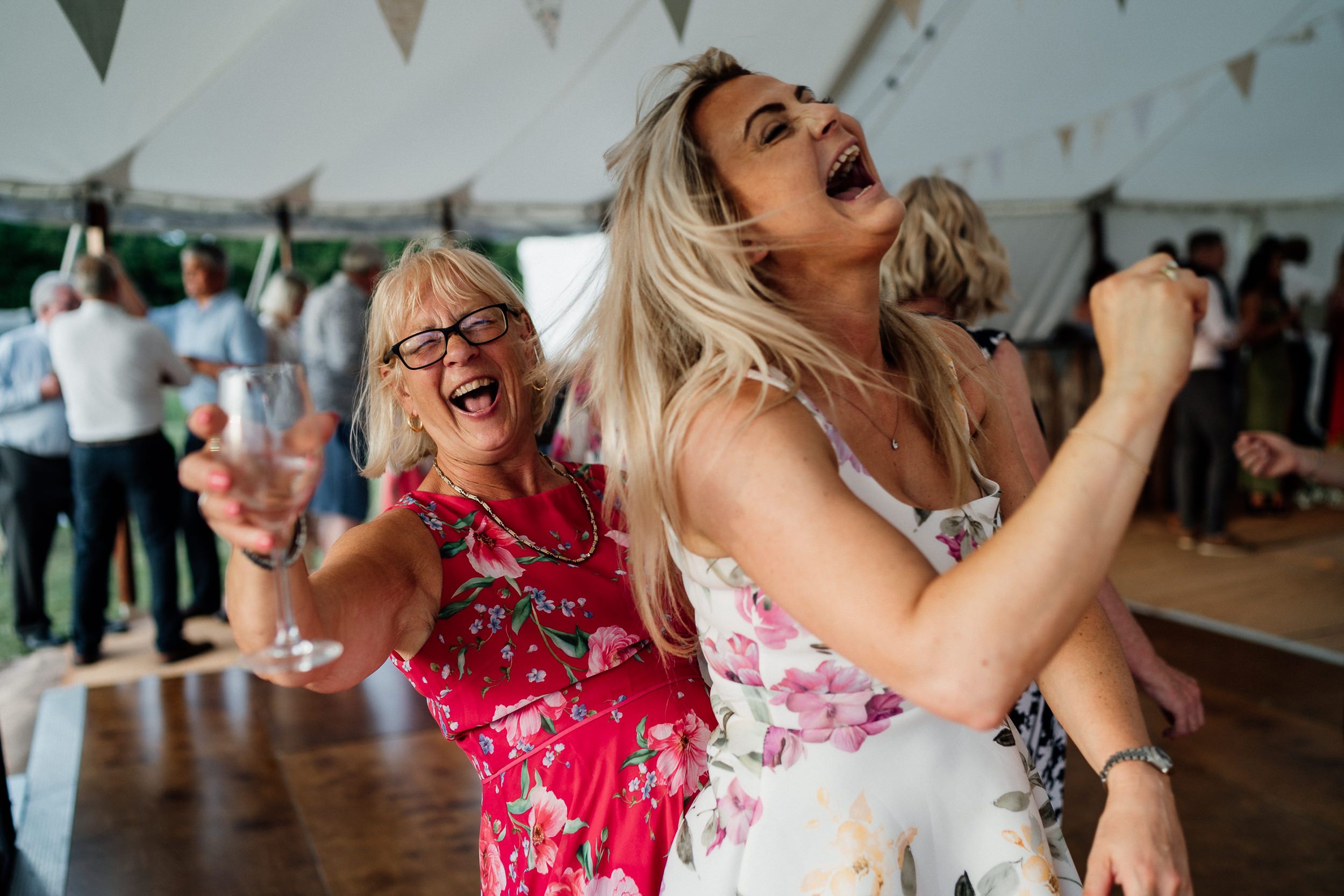 Wedding guests dance enthusiastically during a celebration in a tent with festive bunting decorations.