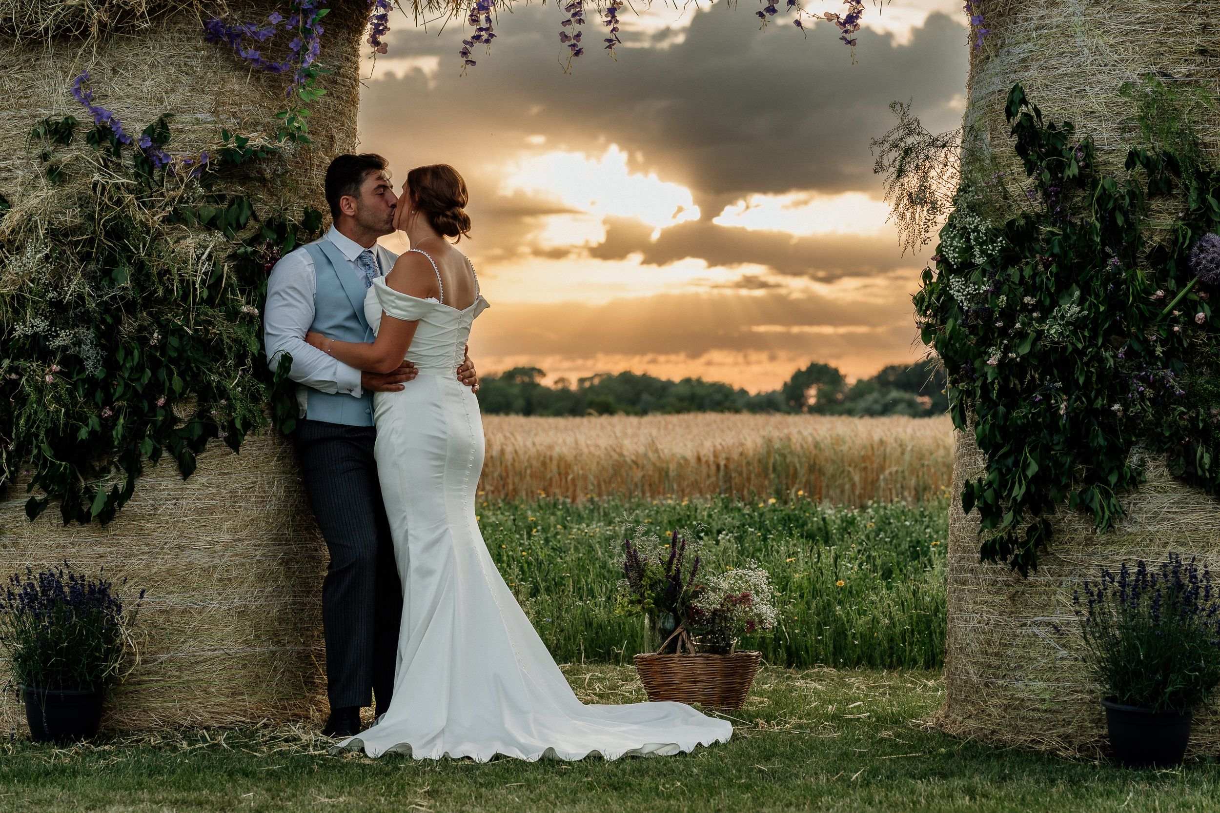 A couple embraces in an elegant wedding photo at sunset, framed by a stone archway with flowering vines.