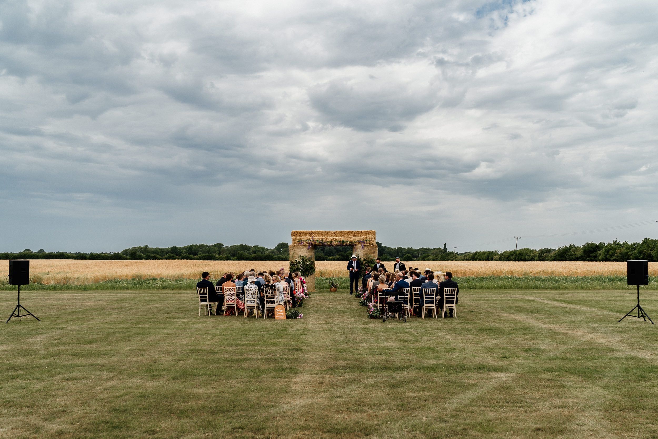 Outdoor wedding ceremony setup on a grassy field with wooden arch under dramatic cloudy sky.