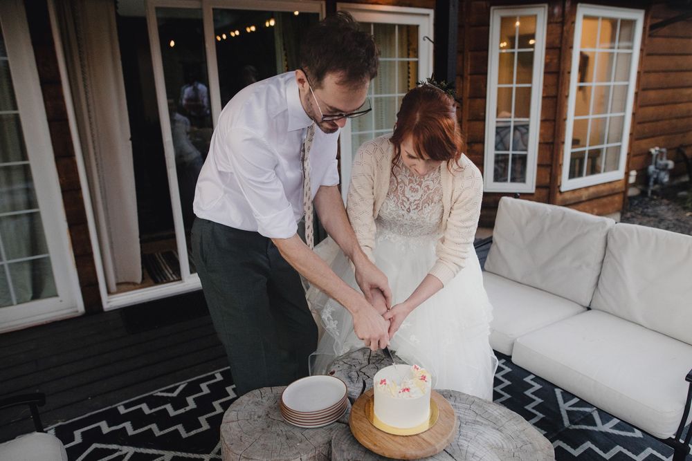 Couple in elegant wedding attire cuts their white cake at rustic outdoor reception venue.