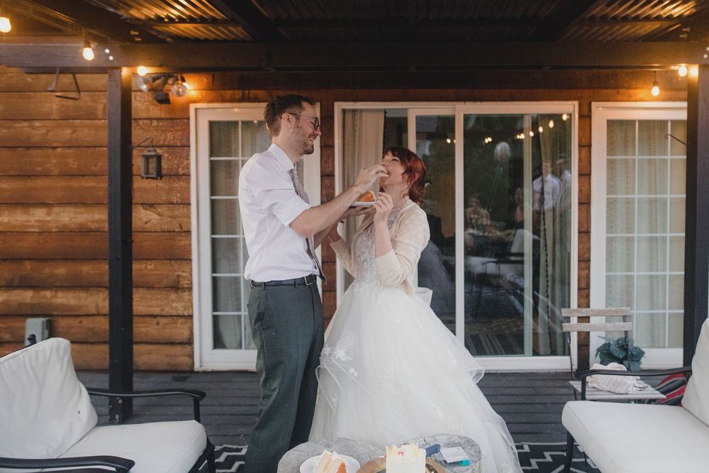 A bride and groom share a playful cake-feeding moment on their wedding day on a rustic cabin porch with string lights.