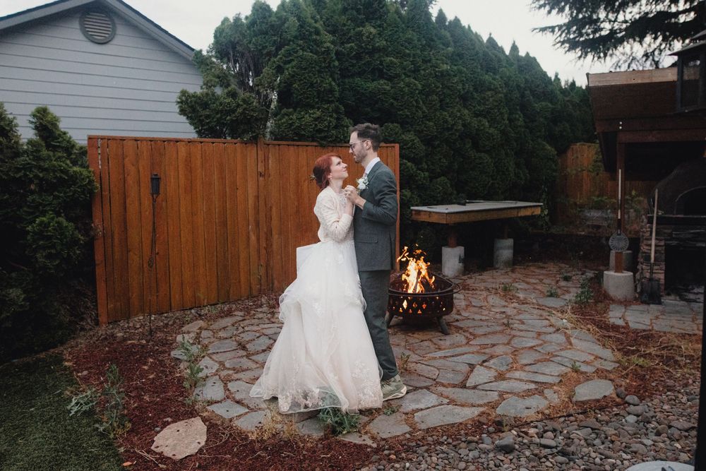 A couple dances together on a stone patio with a wooden fence and firepit glowing in the background.