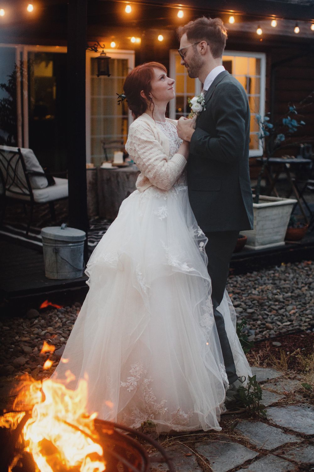 A couple shares a romantic moment by the fire pit at their outdoor evening wedding reception with string lights overhead.