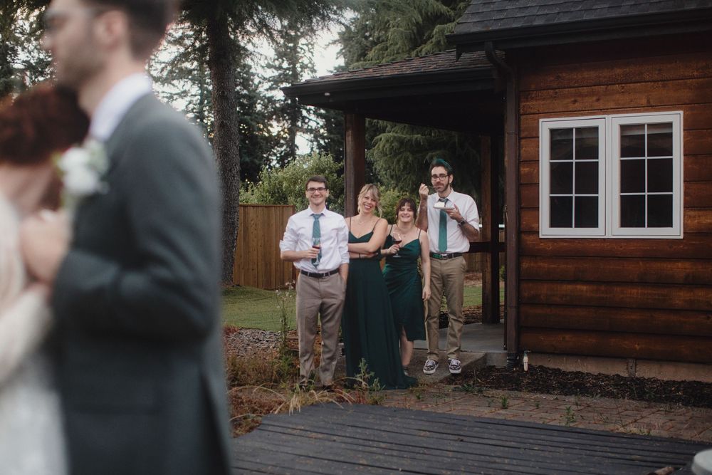 Wedding guests gather outside a rustic wooden cabin venue with dark stained siding and white trim windows.