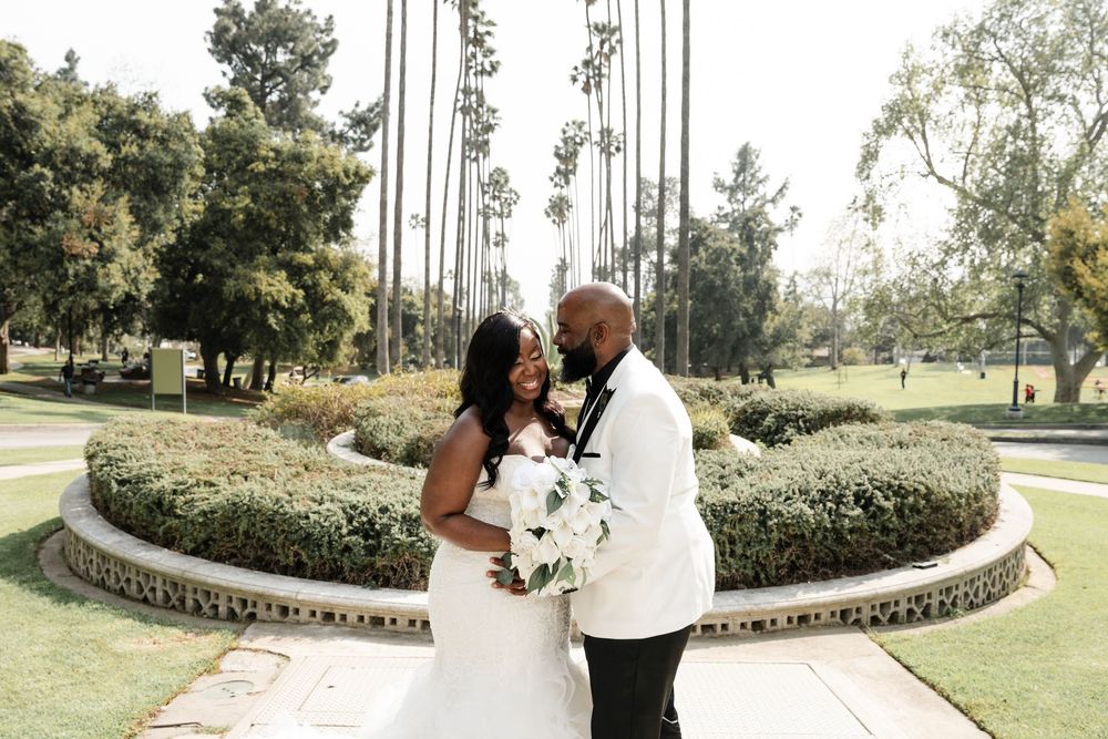 A couple shares romantic embrace at their outdoor wedding ceremony amidst landscaped gardens.