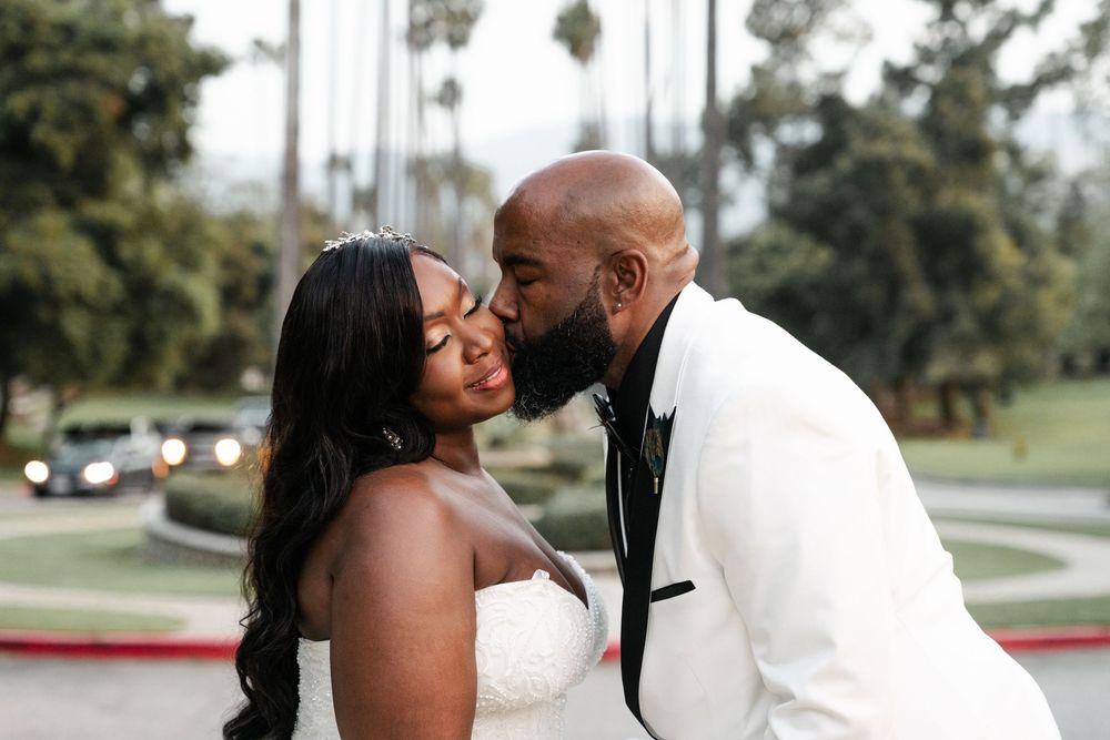 A loving couple shares an intimate moment during their wedding day wearing white formal attire at an outdoor venue.