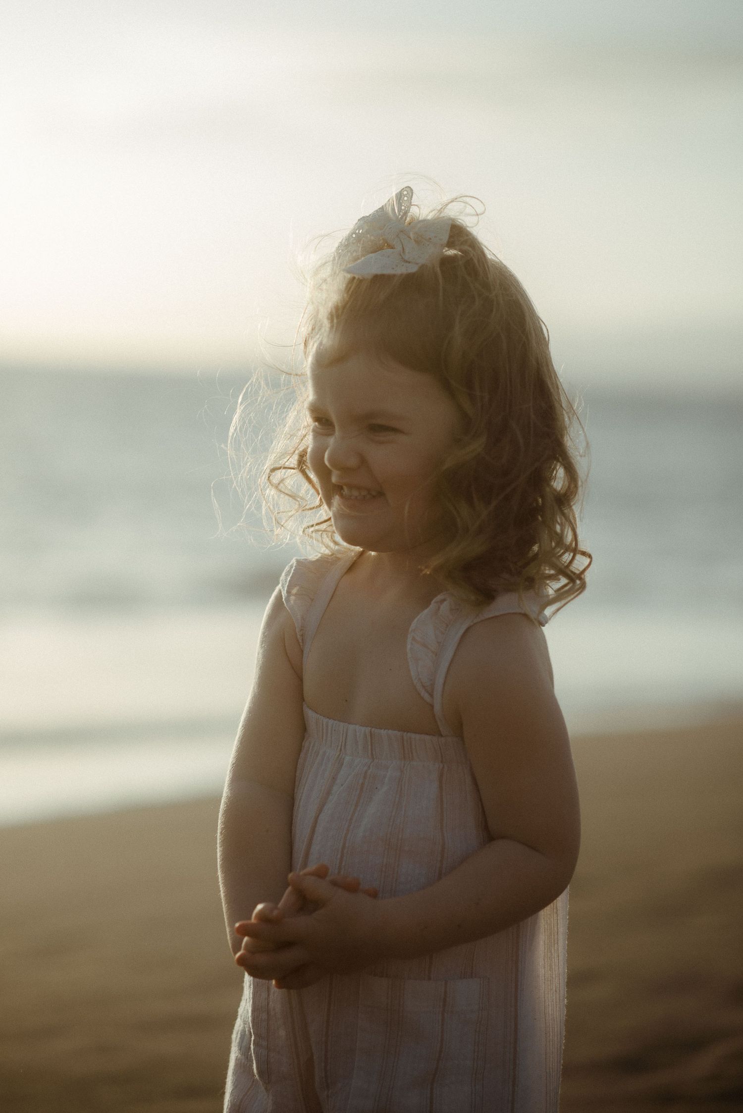 Young child in a white dress laughs joyfully on a sun-lit beach during golden hour. Tenerife Wedding Photographer at Playa del Duque, Adeje.