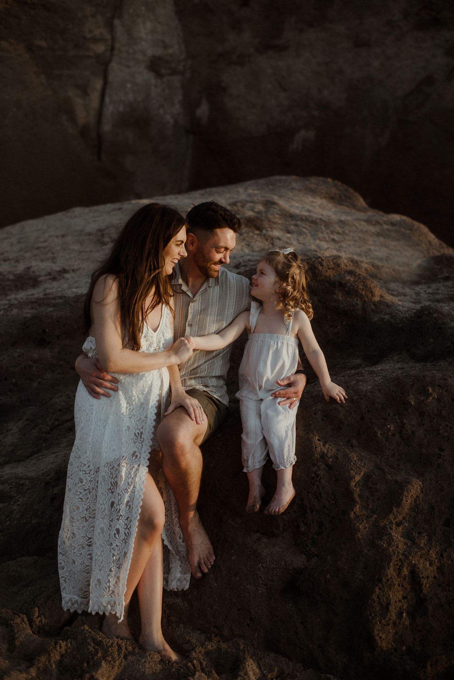 A family wearing white sits together on coastal rocks during golden hour. Tenerife Wedding Photographer at Playa del Duque, Adeje.