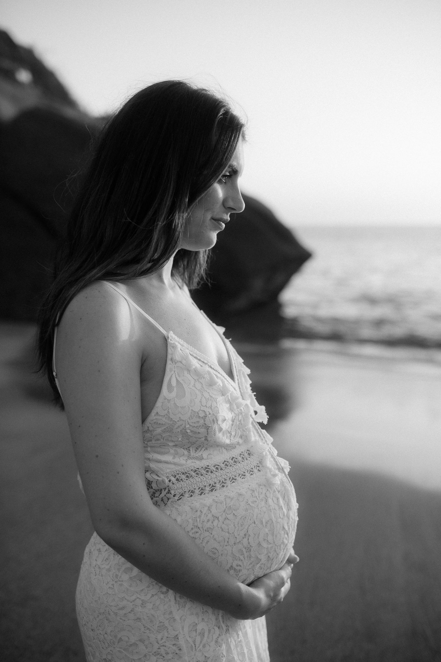 An expectant mother in a white lace dress poses on a beach at sunset. Tenerife Wedding Photographer at Playa del Duque, Adeje.