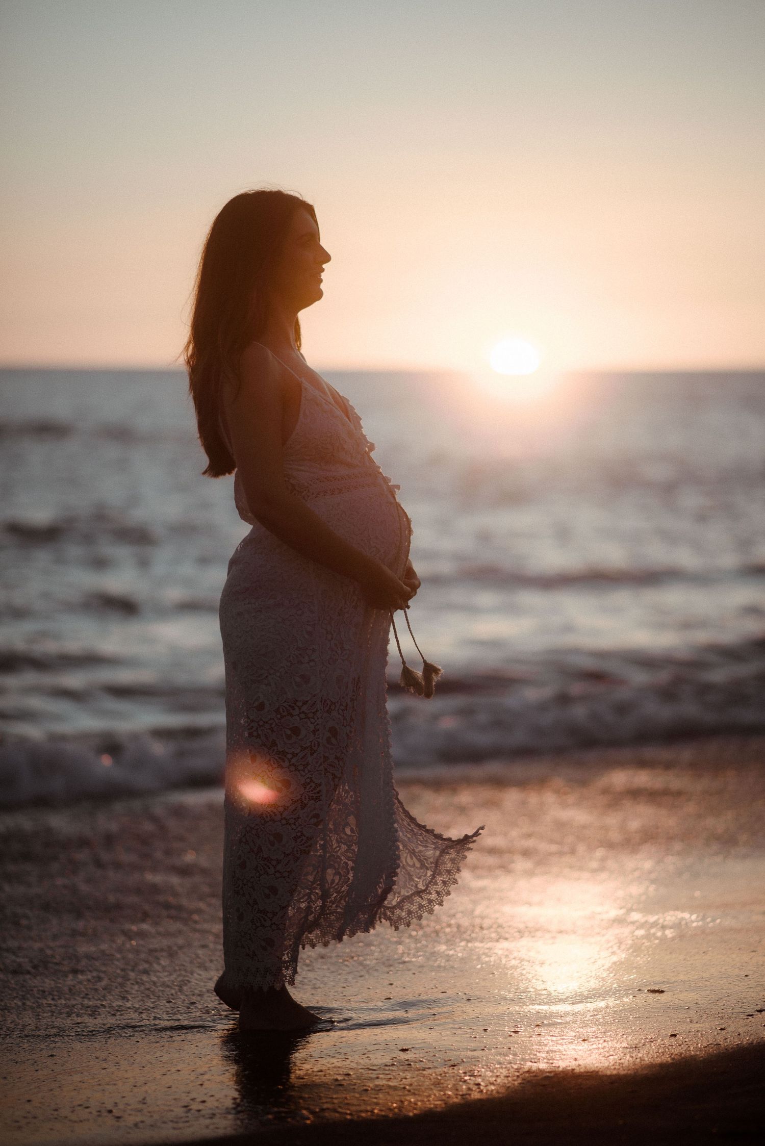 Silhouette of pregnant woman in lace dress watching sunset on beach. Tenerife Wedding Photographer at Playa del Duque, Adeje.