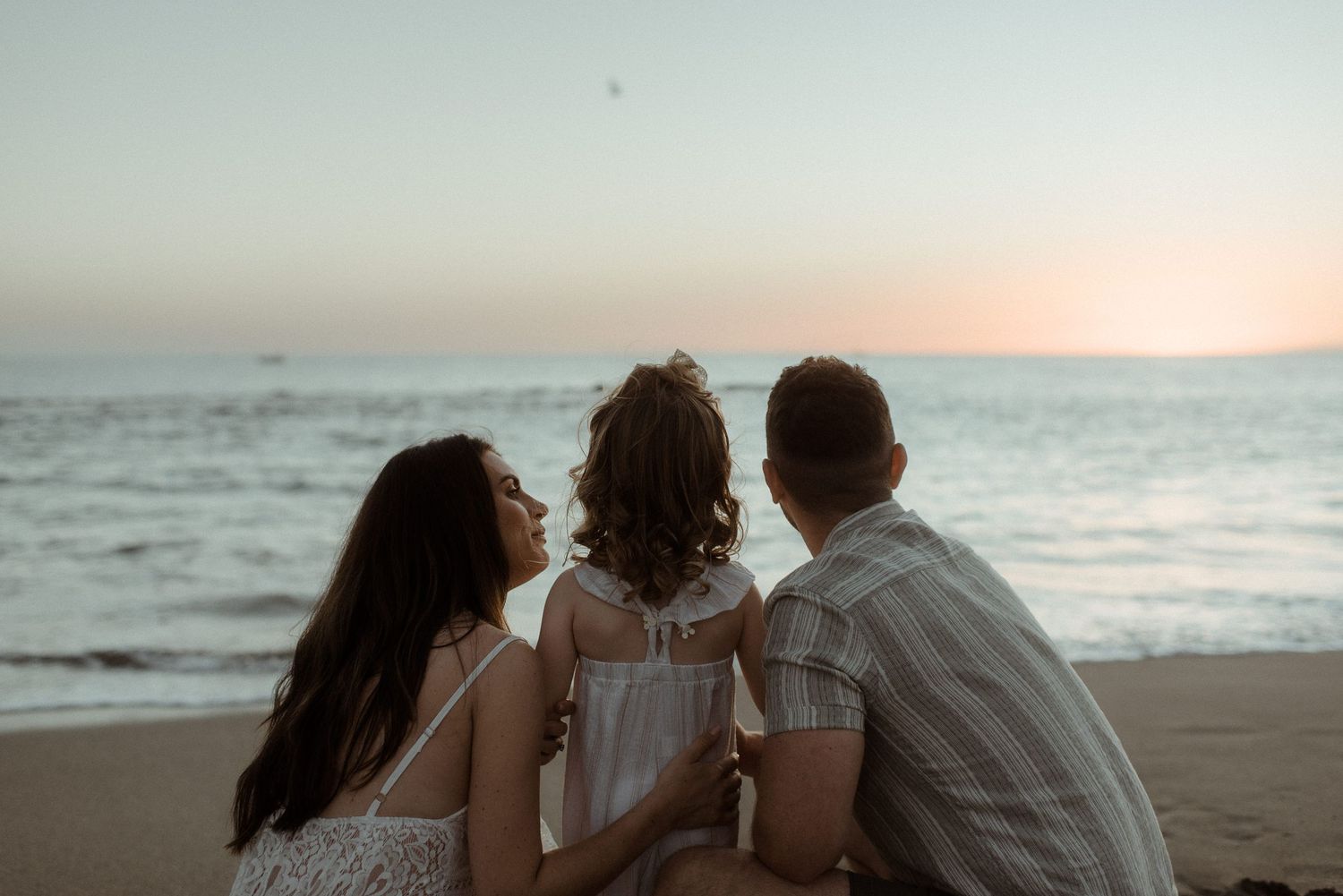 Family of three sits on beach watching sunset together. Tenerife Wedding Photographer at Playa del Duque, Adeje.