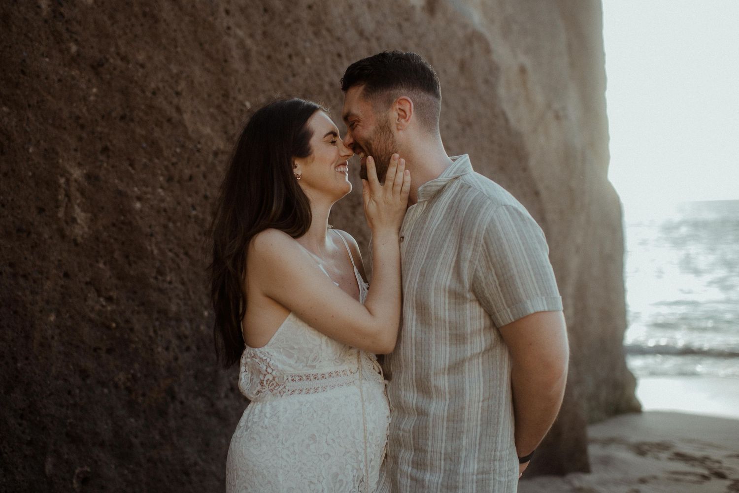 Intimate beach portrait of an expectant couple embracing against a rocky wall at sunset. Tenerife Wedding Photographer at Playa del Duque