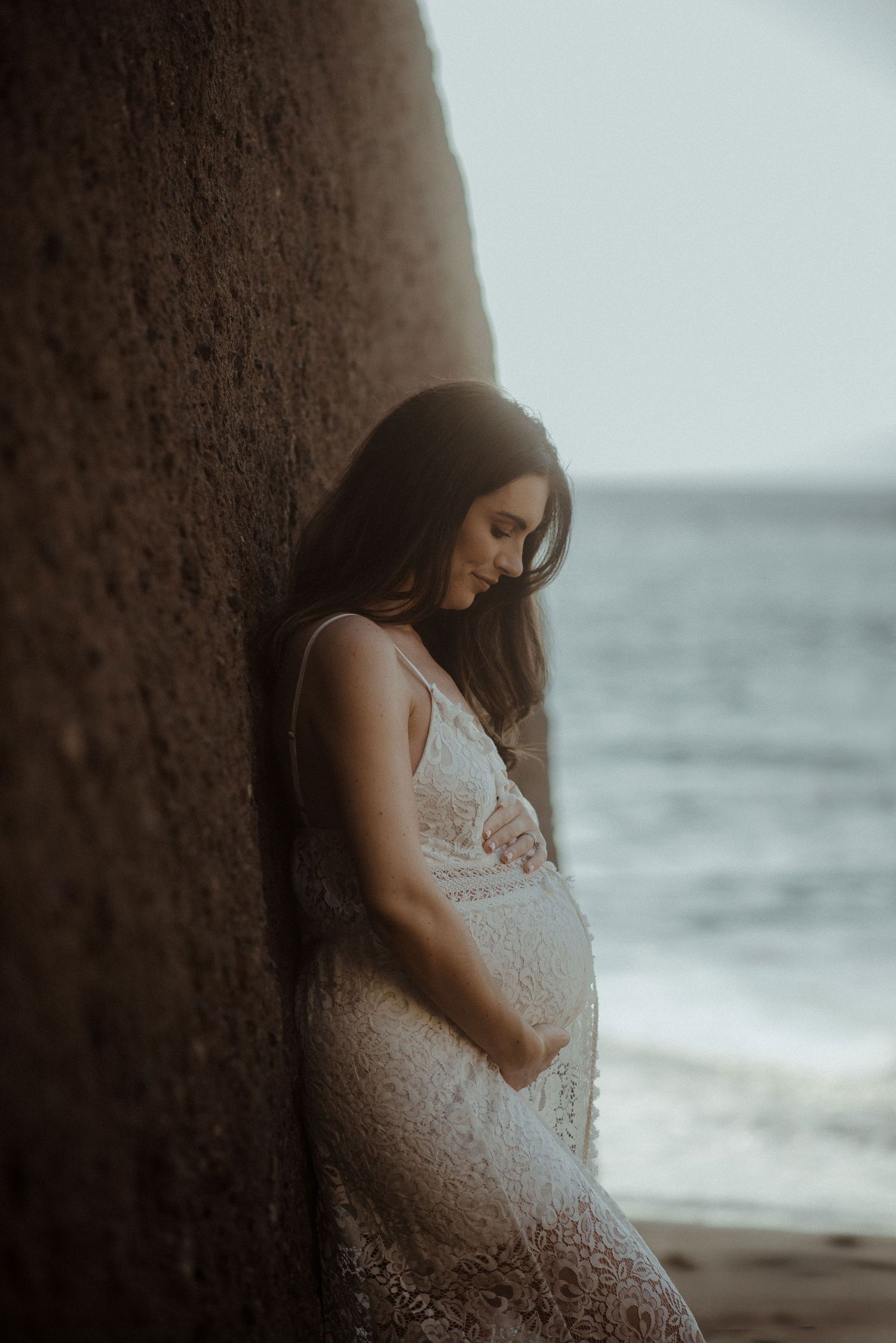 Moody maternity photo of a woman in white lace dress leaning against a cliff wall by the ocean. Tenerife Wedding Photographer