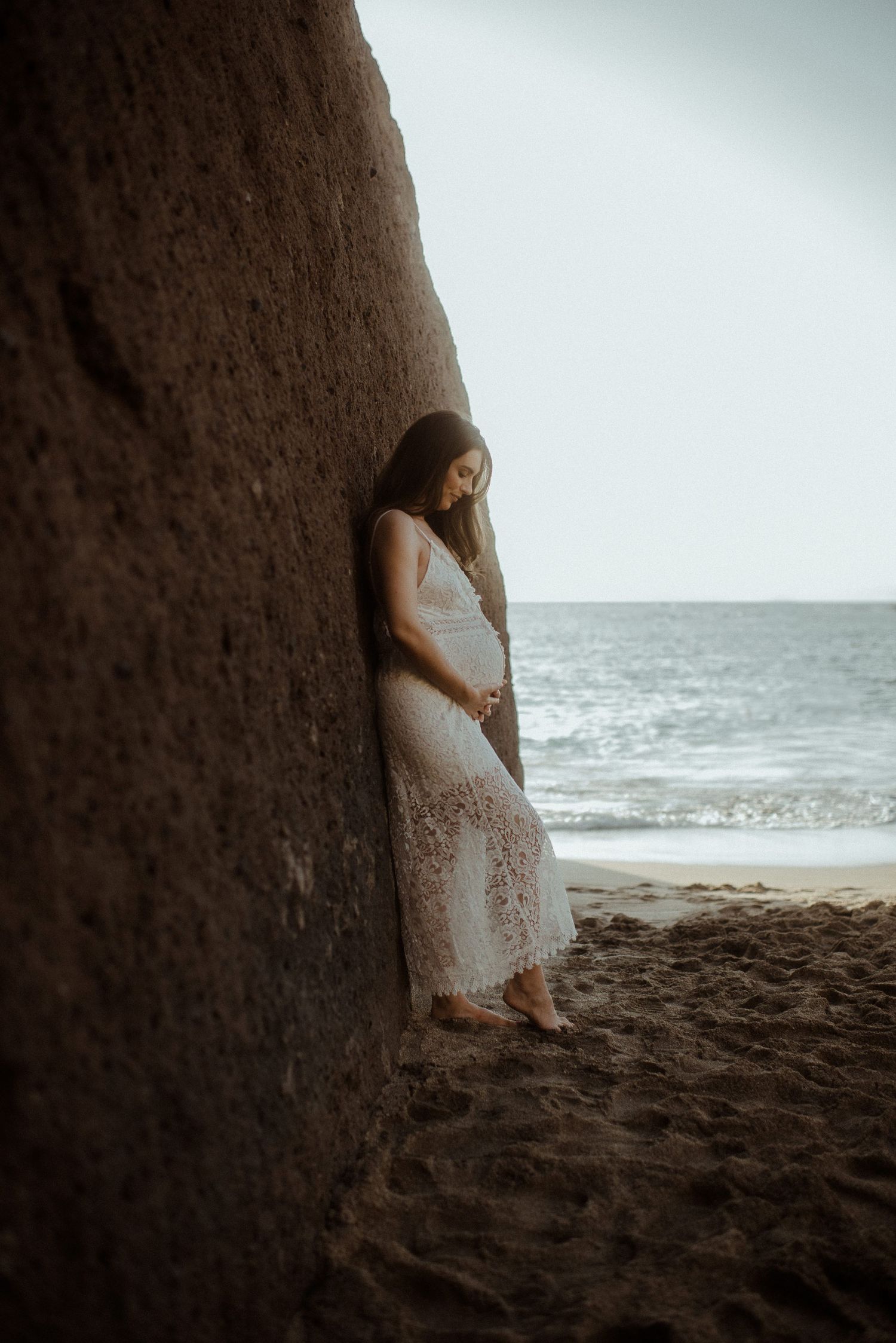 Pregnant woman in white dress standing against rocky cliff with ocean waves in background. Tenerife Wedding Photographer at Playa del Duque