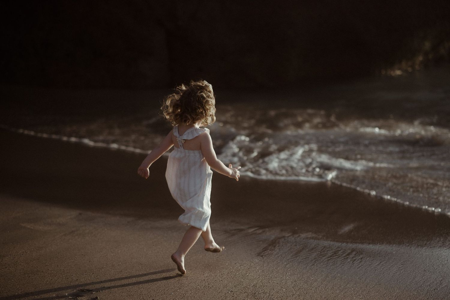 Child in white dress running freely along ocean shore at sunset. Tenerife Wedding Photographer at Playa del Duque, Adeje.