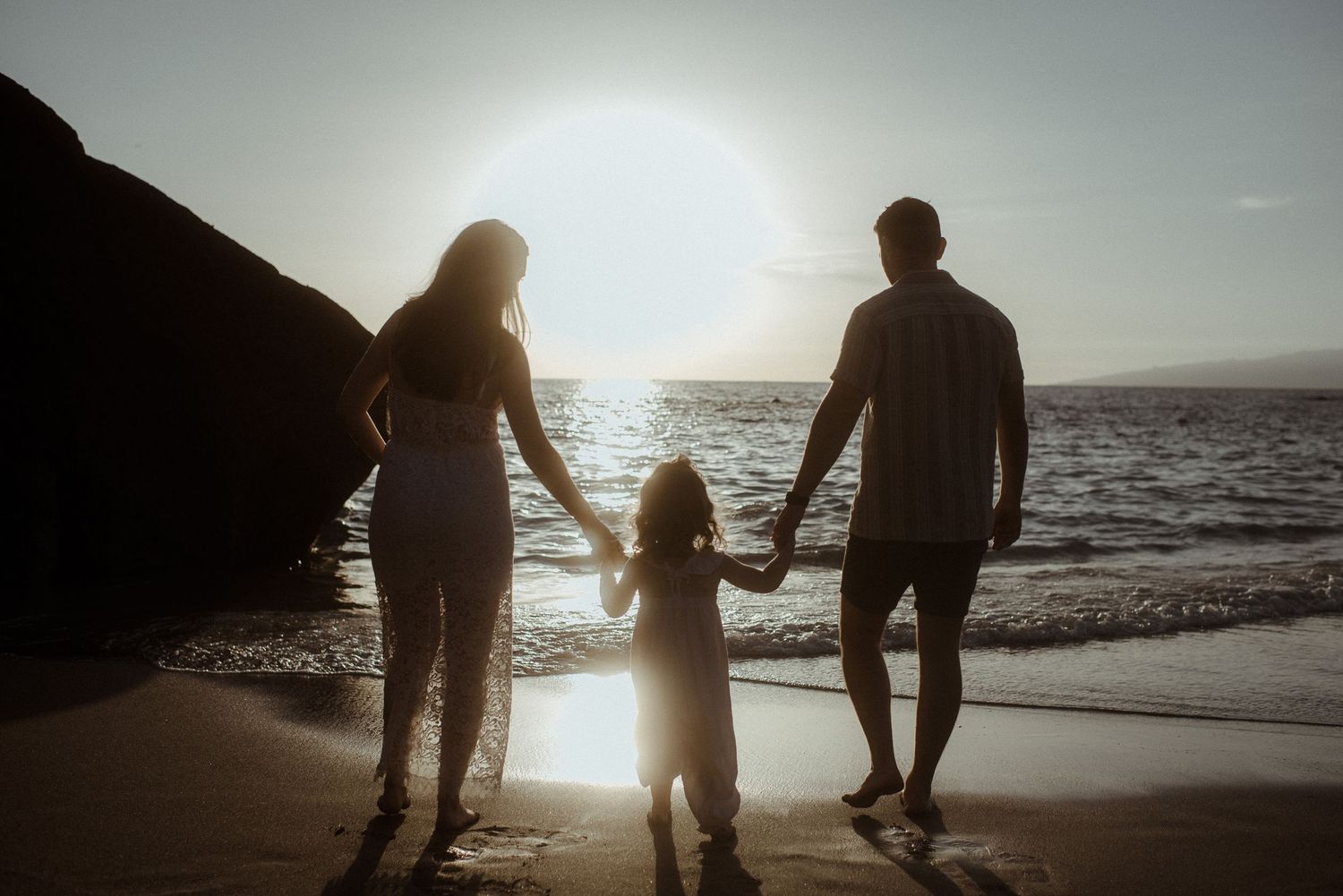A silhouetted family of three holds hands while standing on a beach at sunset. Tenerife Wedding Photographer at Playa del Duque, Adeje.