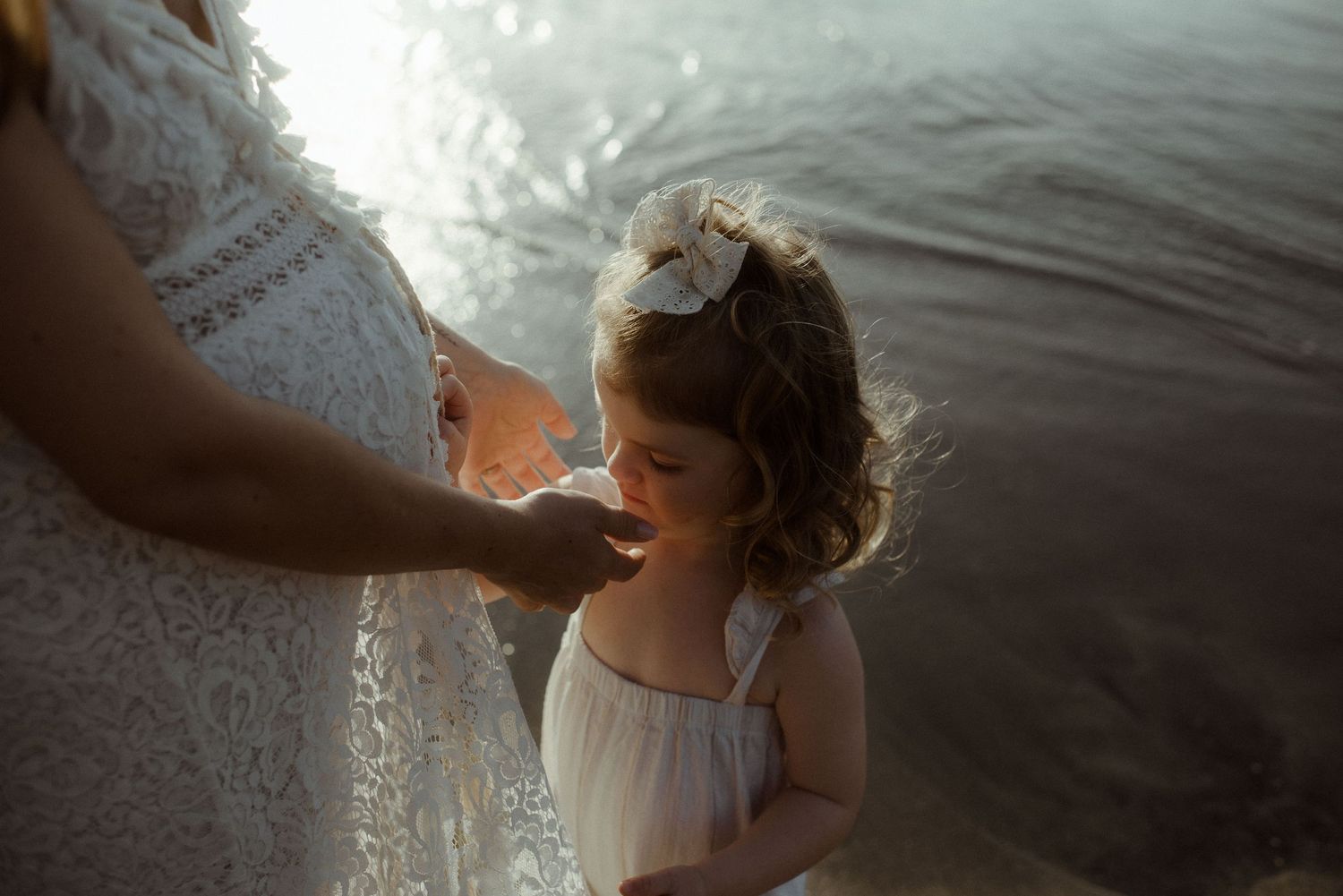A small child in a white dress smiles while standing by the water at sunset. Tenerife Wedding Photographer at Playa del Duque, Adeje.
