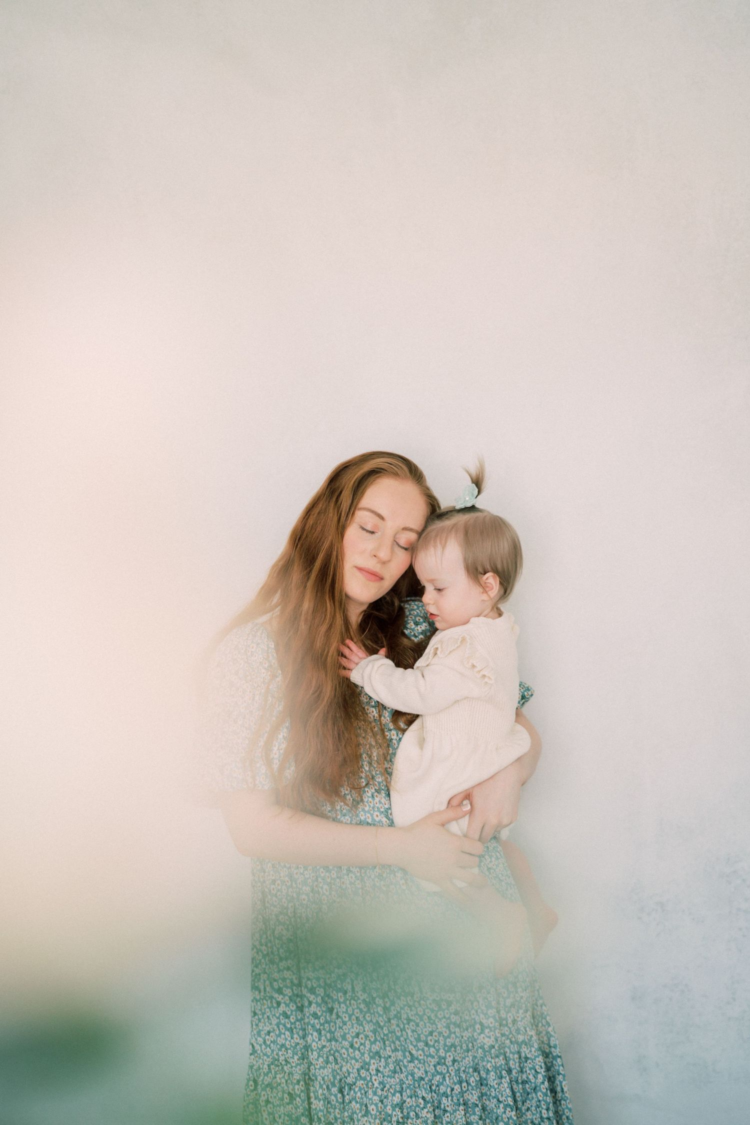 A tender moment captured as a woman in a patterned dress holds a baby in white against a soft neutral background.