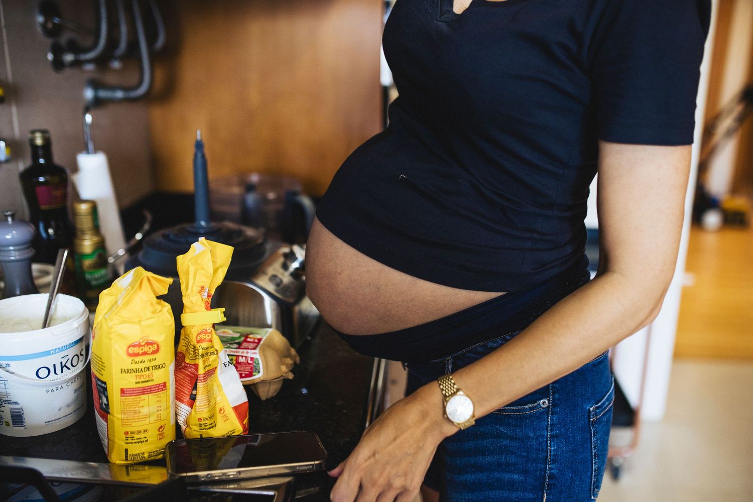 Pregnant person in dark top and jeans stands in kitchen with yellow grocery bags on counter.