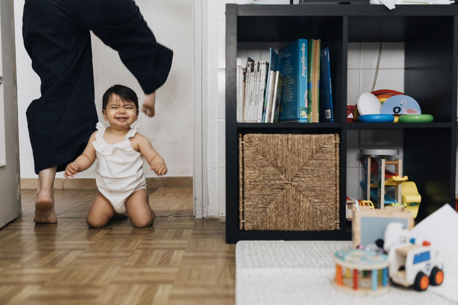 A toddler in a white onesie plays near a black bookshelf filled with books and toys on a wooden floor.