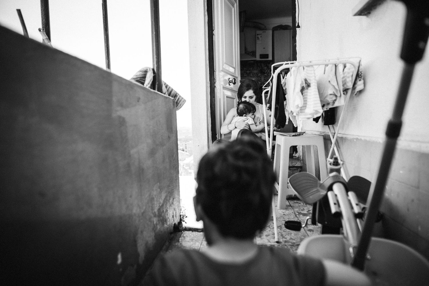 A monochromatic candid shot inside a cluttered room with clothing hanging in the background.