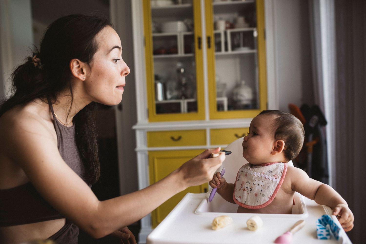 Parent feeds baby in a highchair while sitting at a kitchen table with yellow cabinets in the background.