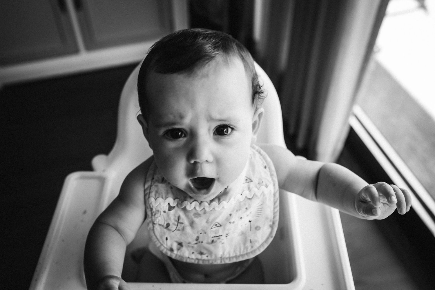 A happy baby with a big smile sitting in a high chair by the window, captured in black and white.