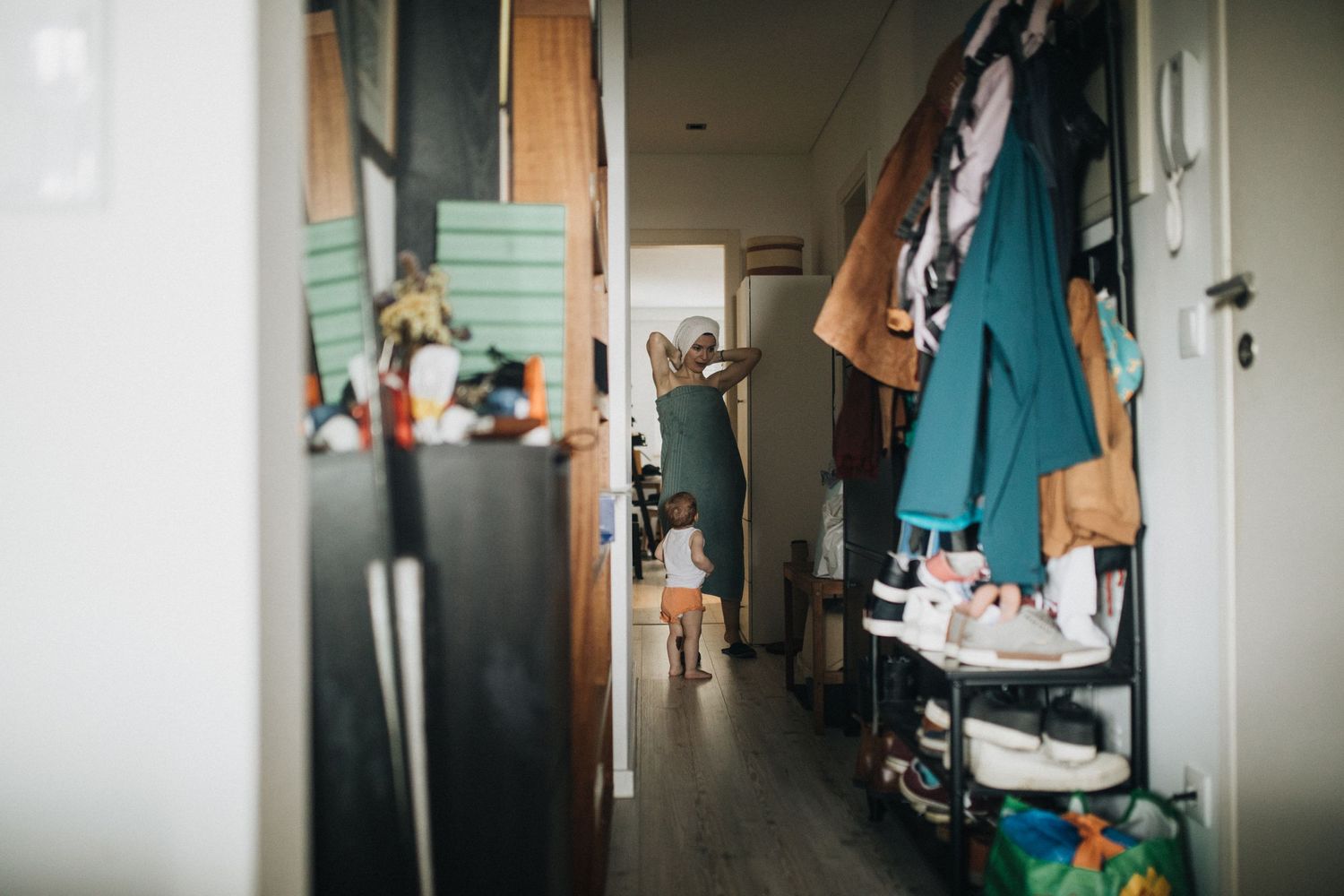 Interior view of a cluttered residential hallway with coats and shoes along one wall and soft natural lighting.