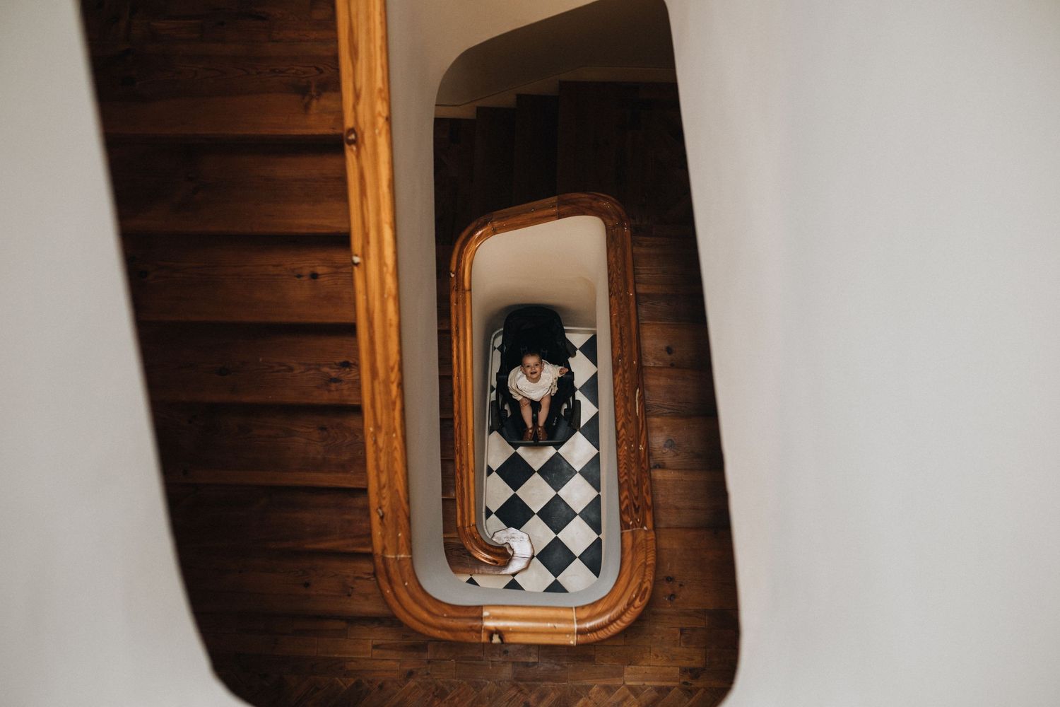 Looking down a spiral wooden staircase with geometric black and white checkered floor pattern visible at the bottom.