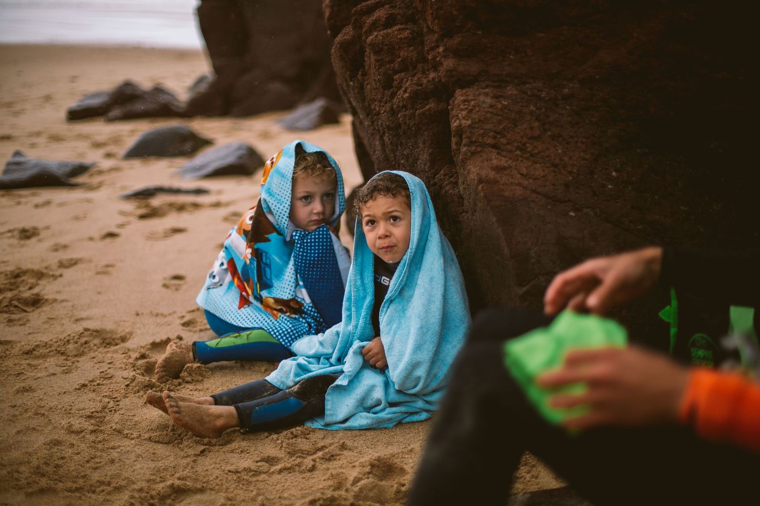 Two figures wrapped in bright blue blankets sit on a sandy beach near rocky cliffs at dusk.