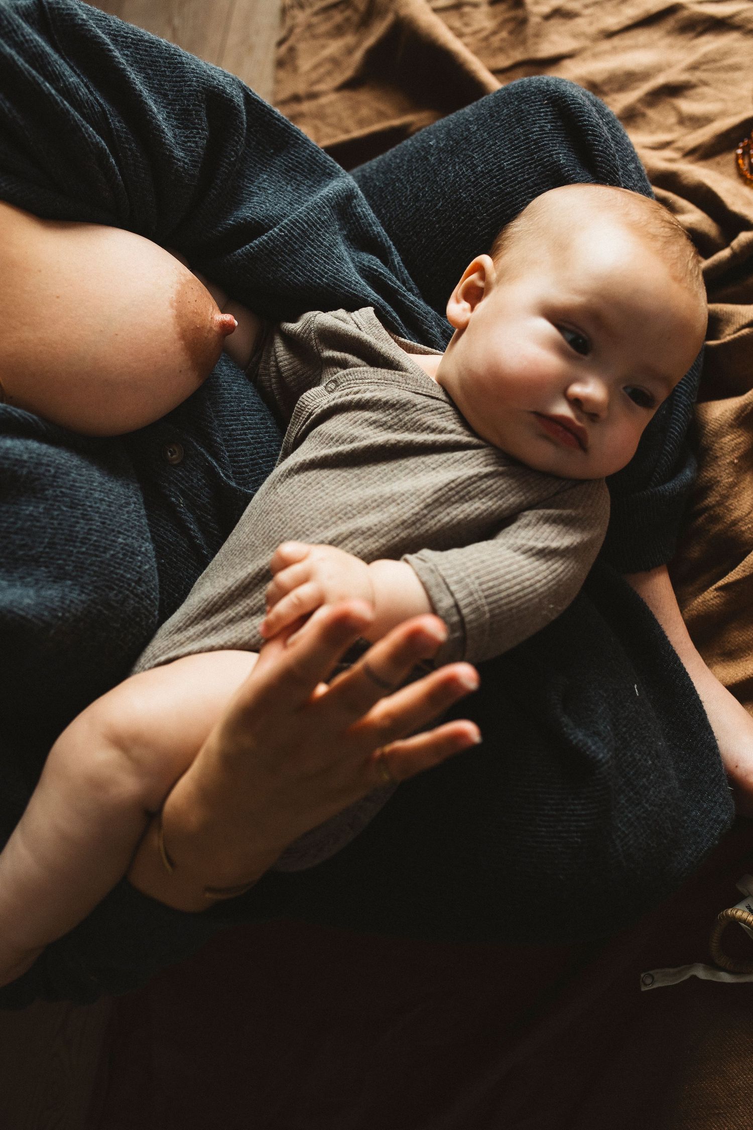 A baby wearing a gray onesie lies peacefully on dark fabric with a hand gently resting on their tummy.