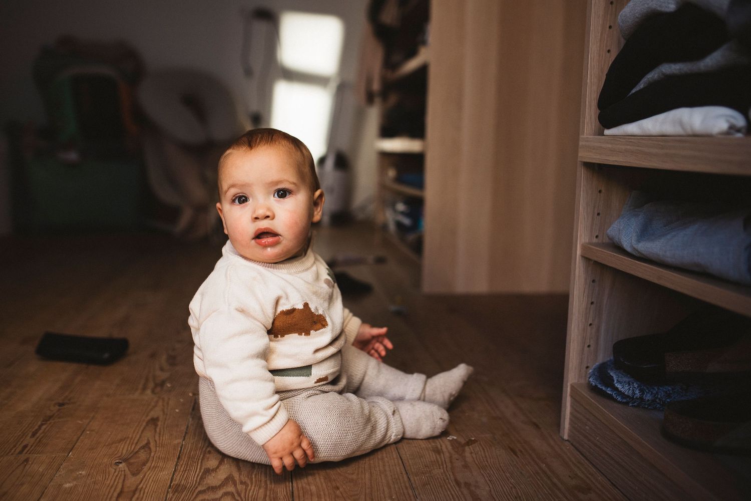 Baby in white outfit with brown bear design sitting on hardwood floor by closet shelf.